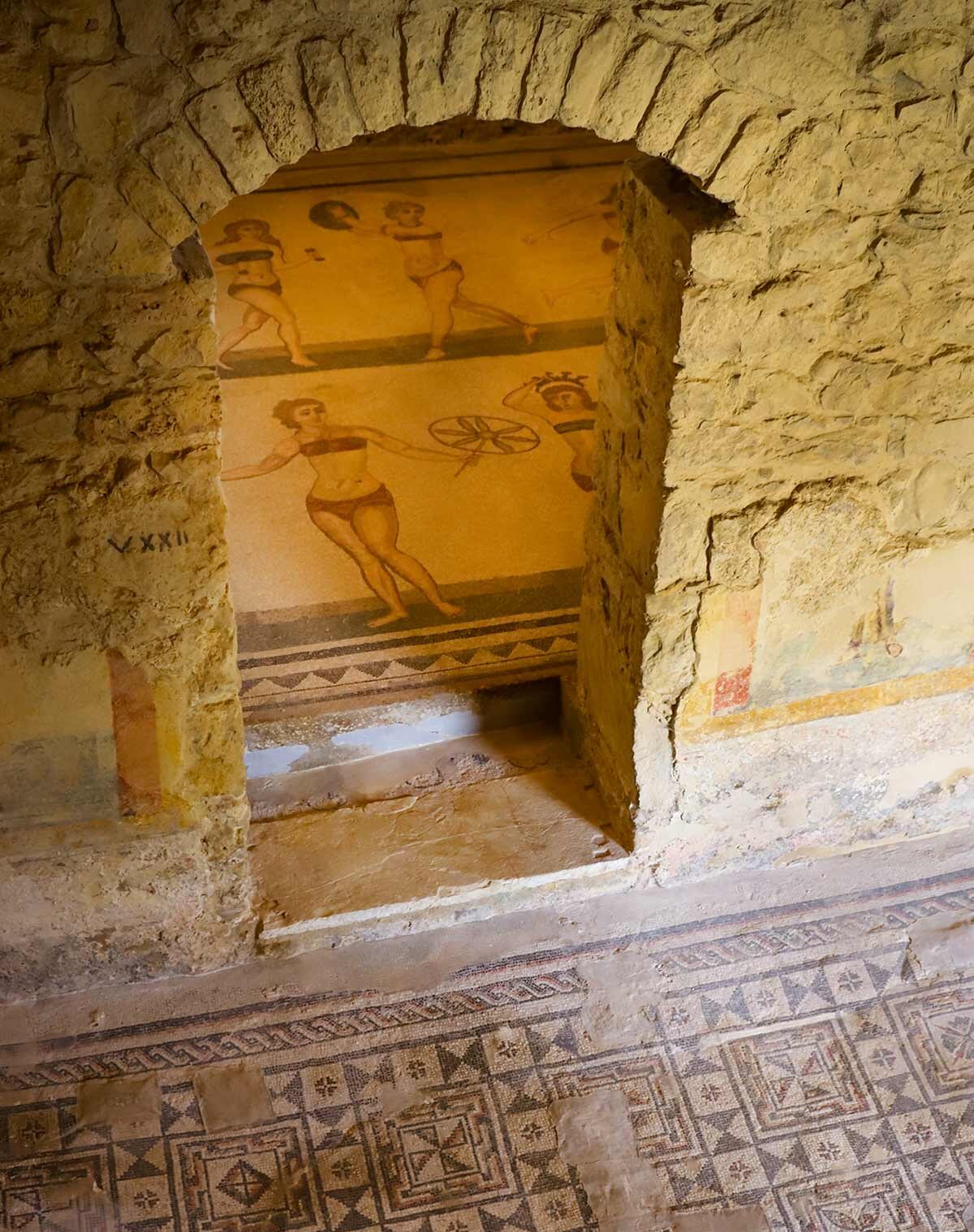 Römische Mosaikboden in der Villa Romana del Casale zeigt Frauen in antiken Bikinis, umgeben von Steinmauern.