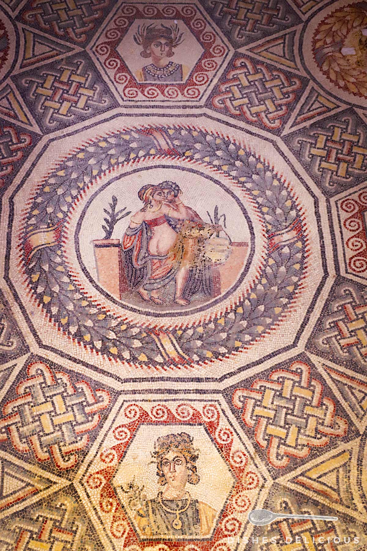 Antikes Bodenmosaik mit zentralem Medaillon Frau und Mann beim Sex, umgeben von geometrischen Mustern und Lorbeerkranz.