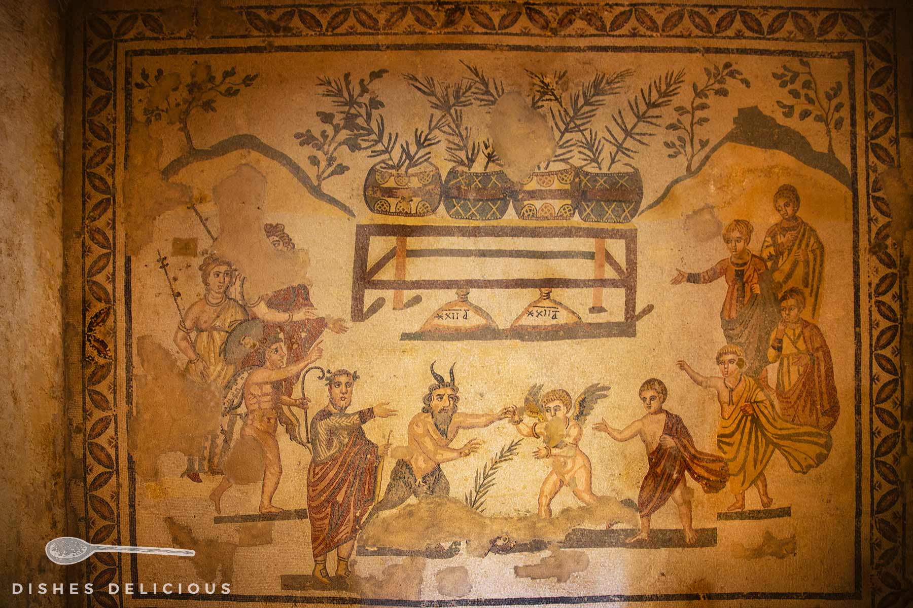 Römisches Mosaik zeigt einen Wettkampf zwischen Eros und Pan, umgeben von Figuren mit antiker Kleidung und Pflanzen in Vasen auf einem Tisch.