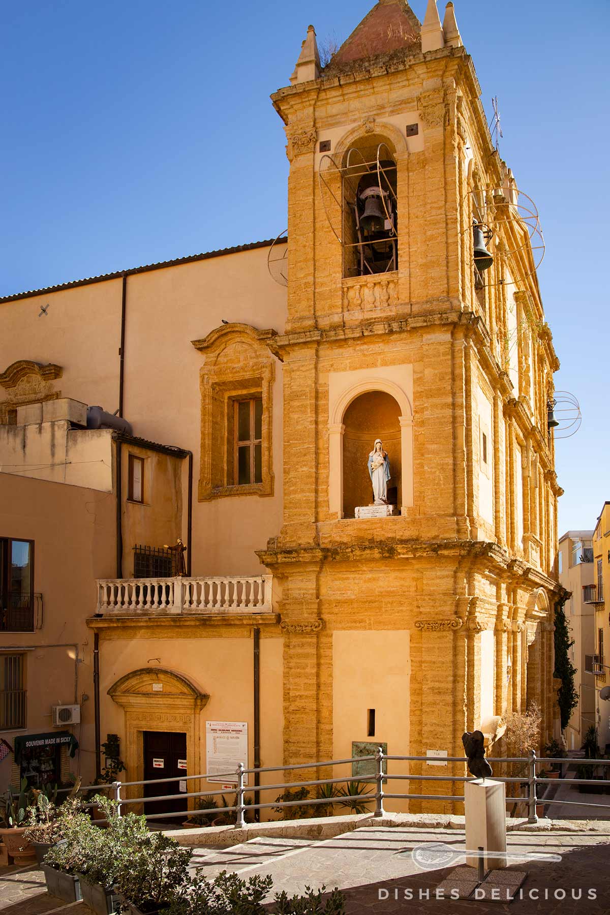 Eine historische Basilica della Immacolata aus gelbem Stein mit einem Glockenturm, einer Statue in einer Nische und einem kleinen Platz davor, unter einem strahlend blauen Himmel.