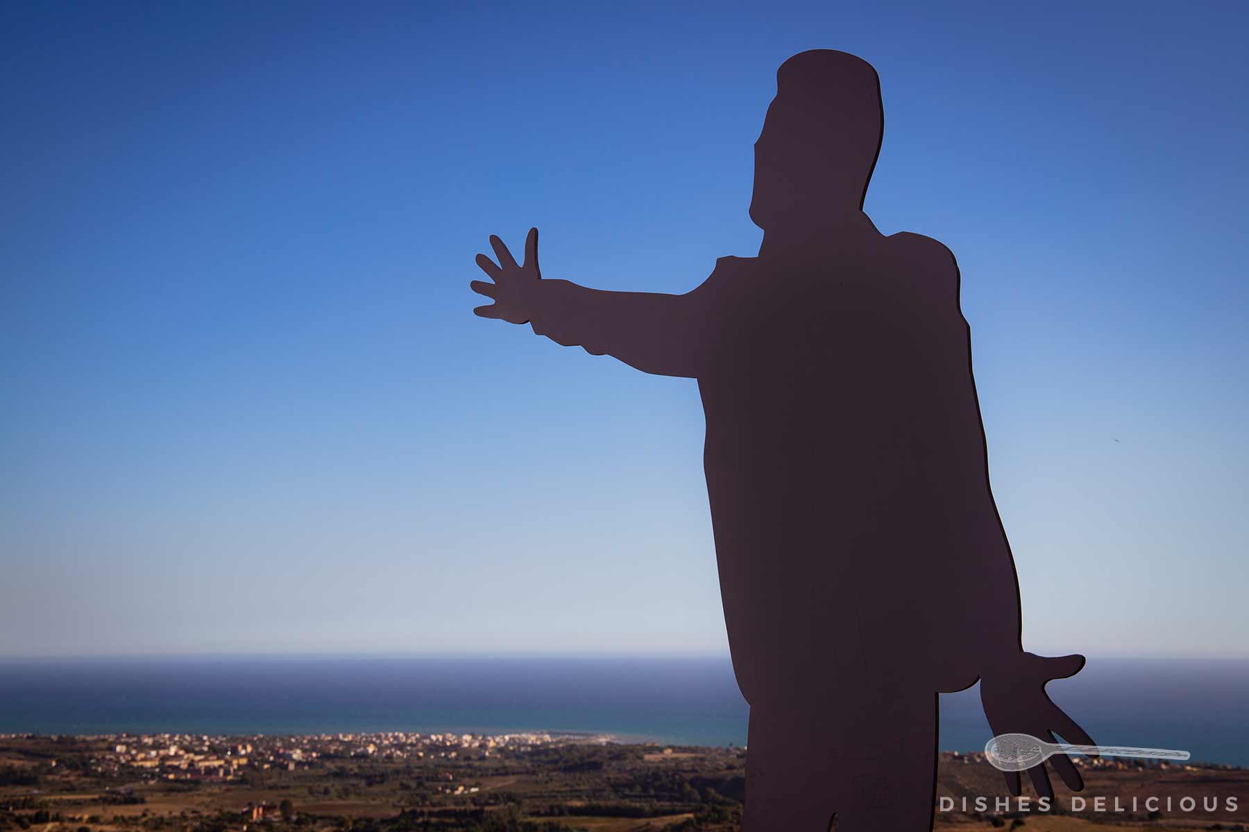 Silhouette einer Statue von Domenico Modugno mit ausgebreiteten Armen vor einem Panorama mit Stadt und Meer am Horizont.