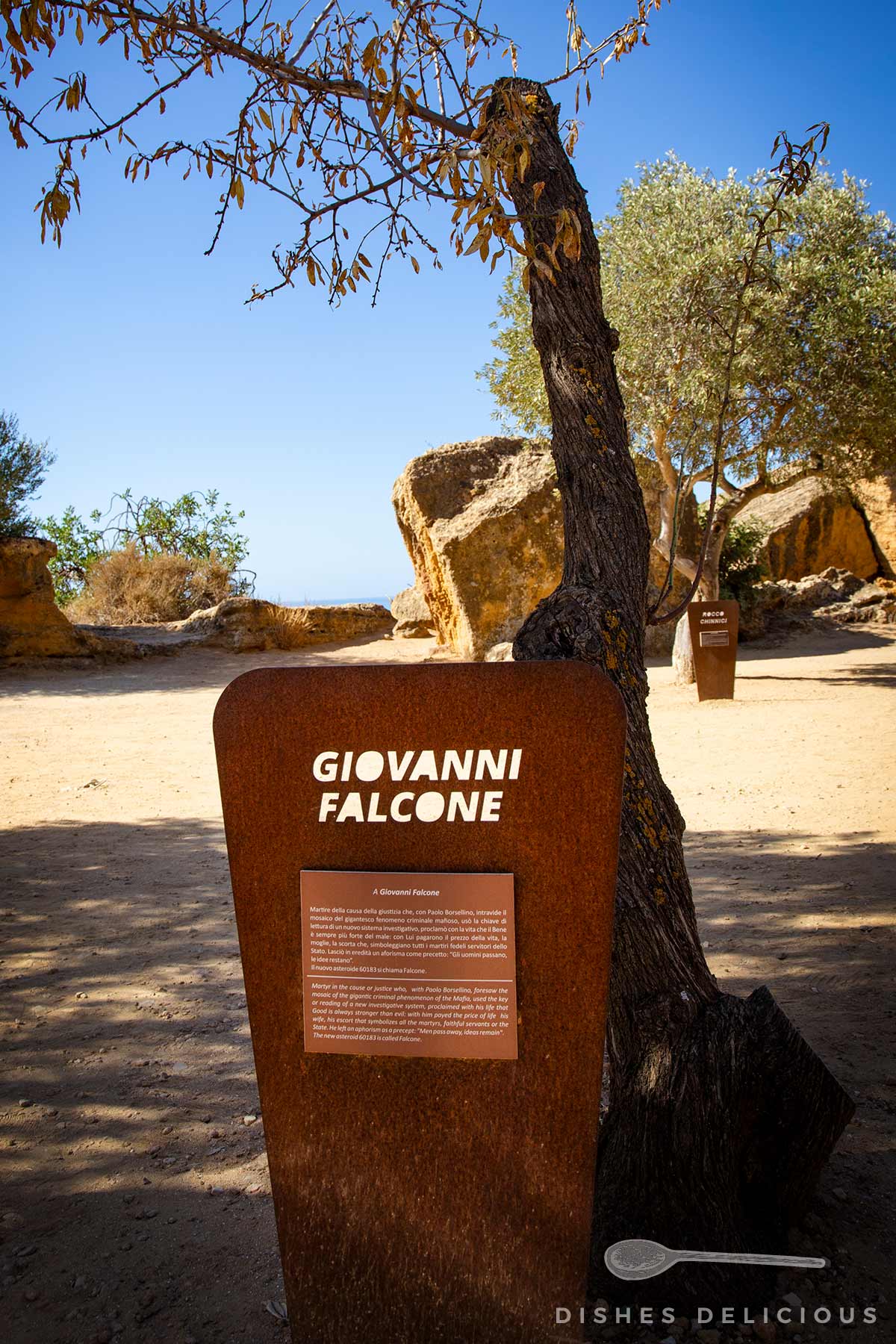 Gedenkstein mit der Aufschrift 'Giovanni Falcone' und Texttafel, im Garten der Gerechten in Sizilien, umgeben von Bäumen und Felsen unter blauem Himmel.