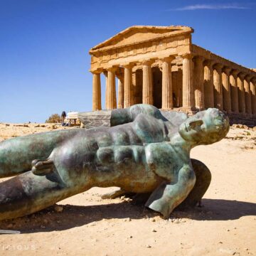 Bronzefigur des liegenden Ikarus vor dem antiken dorischen Concordia-Tempel in Agrigent unter blauem Himmel.