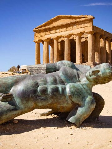 Bronzefigur des liegenden Ikarus vor dem antiken dorischen Concordia-Tempel in Agrigent unter blauem Himmel.