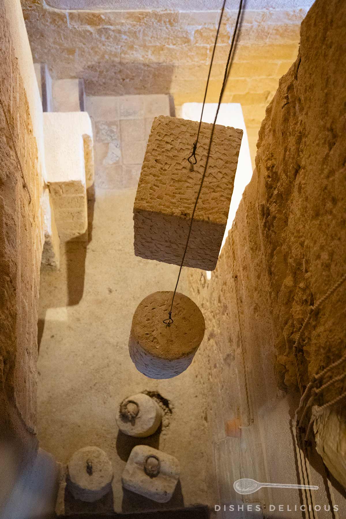 Blick in den Glockenturm des Doms von Agrigento mit zwei hängenden Steinblöcken und mehreren runden Steinen auf dem Boden aus hellem Sandstein.