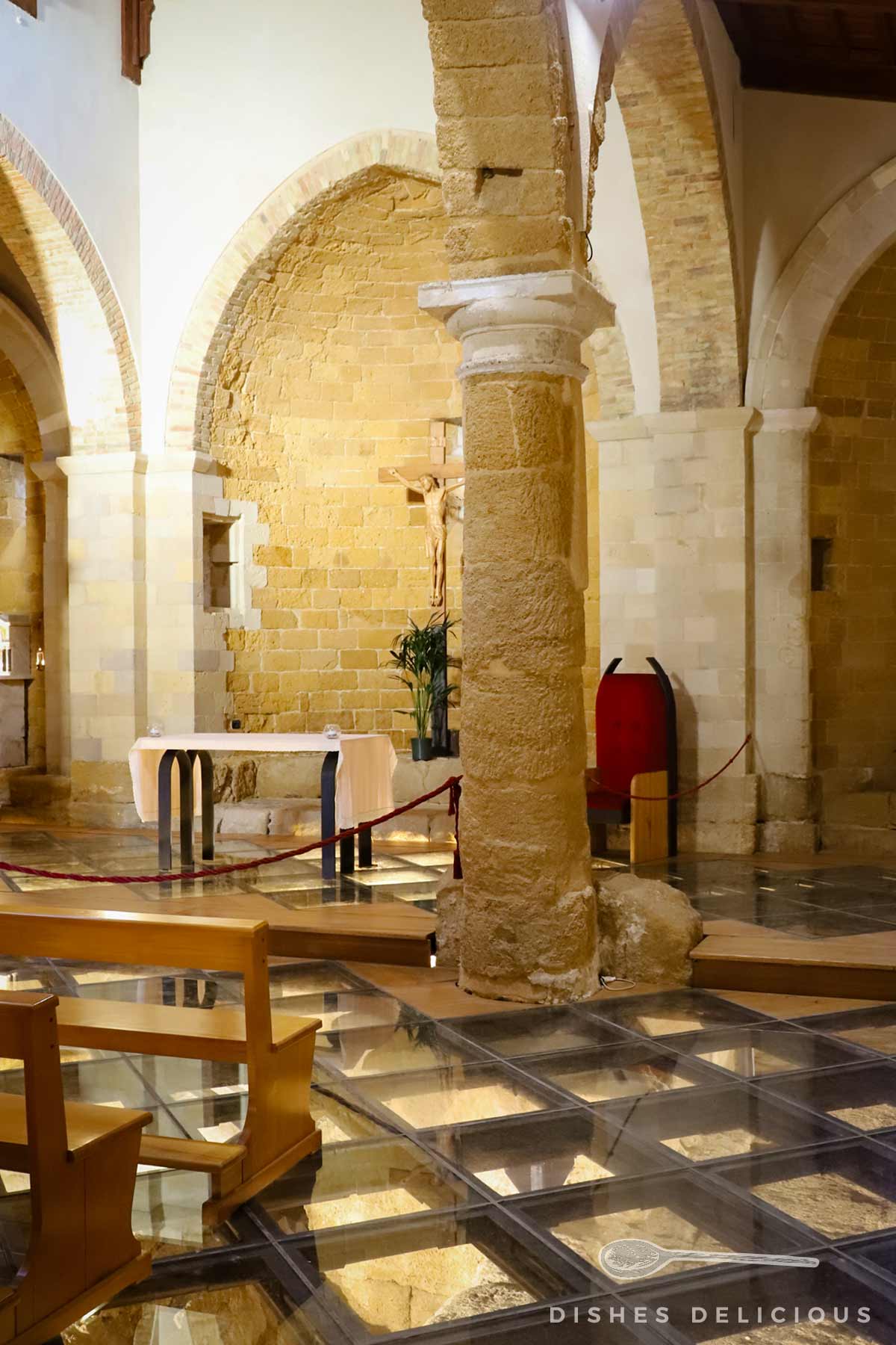 Innenraum der Santa Maria dei Greci in Agrigento mit steinernen Säulen, Glasboden und einem Altar mit Kreuz im Hintergrund.