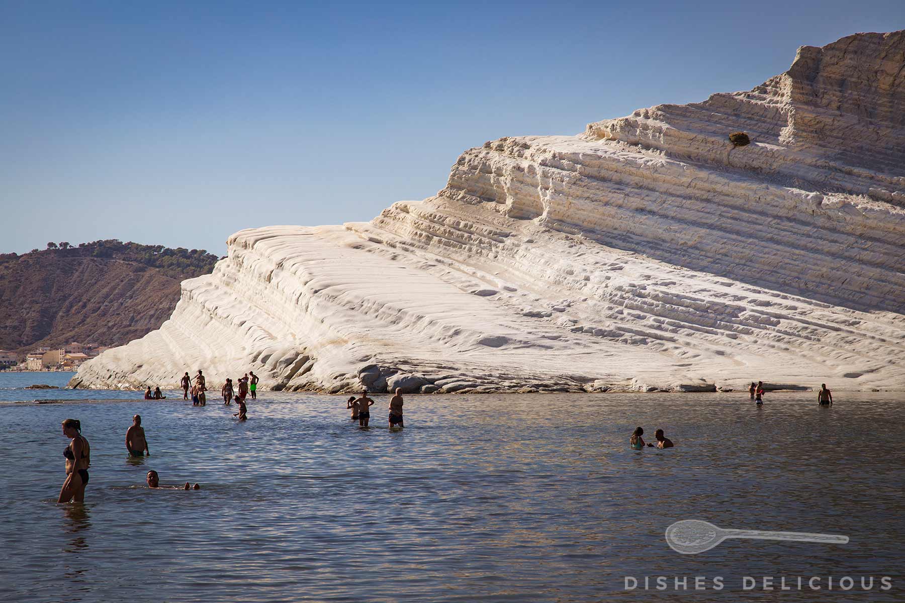 Menschen baden im Meer vor der weißen, terrassenförmigen Kalksteinformation Scala dei Turchi an der Küste Siziliens unter blauem Himmel.