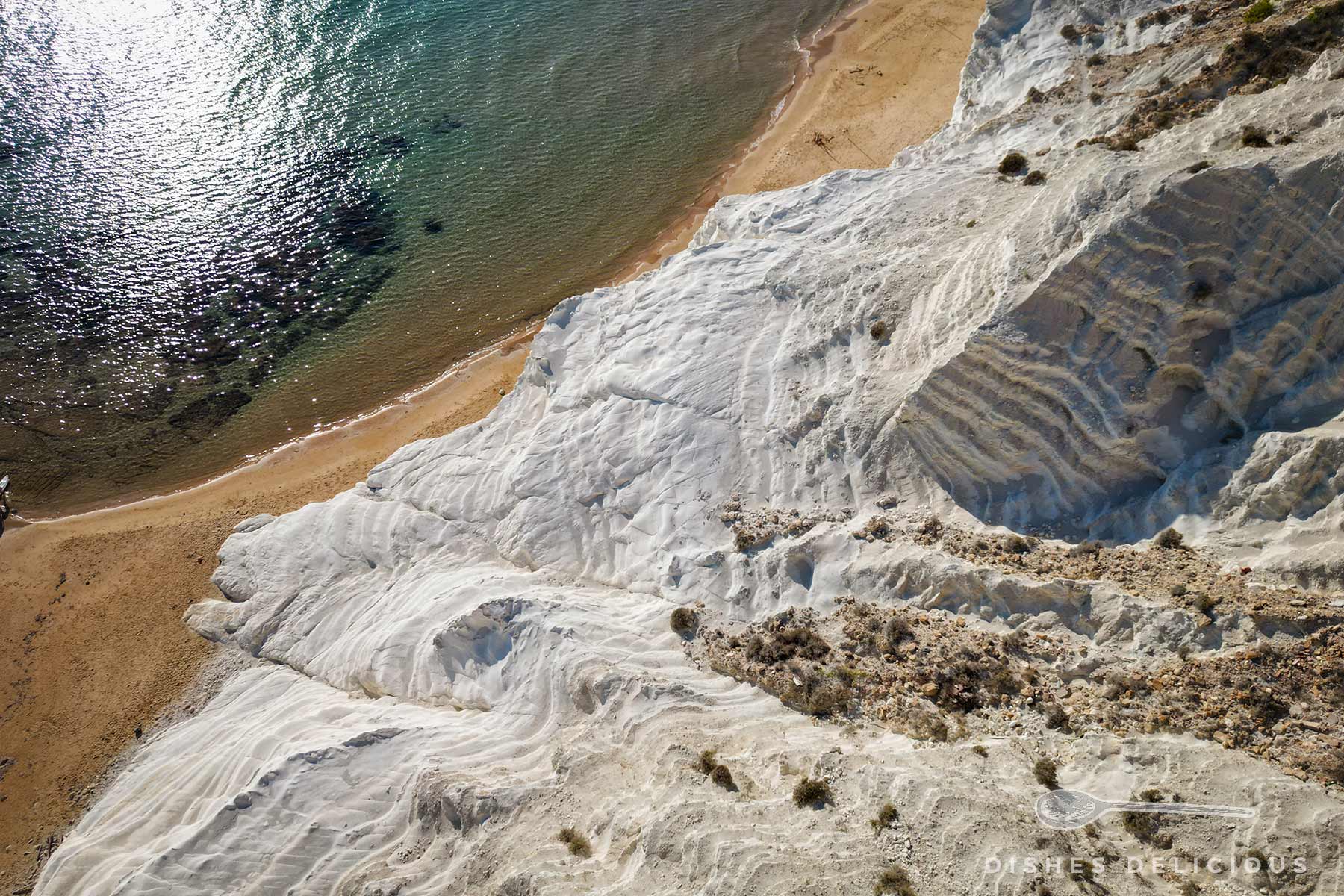 Weiße, terrassenförmige Kalksteinfelsen der Scala dei Turchi neben einem sandigen Strand und ruhigem Meerwasser mit Sonnenreflexionen.