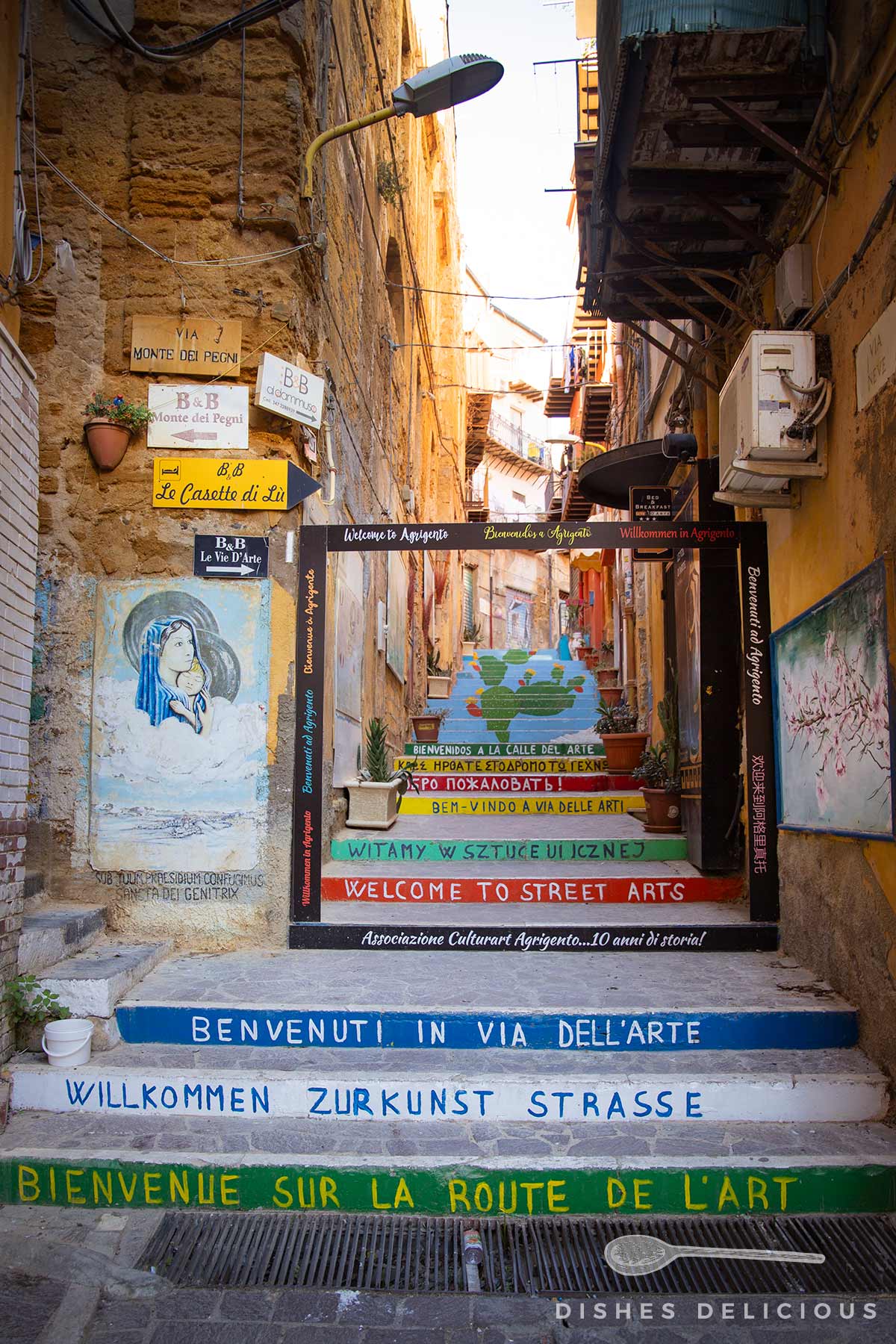 Bunte Treppe mit mehrsprachigen Willkommensgrüßen zu Kunst und Straßenkunst, umrahmt von alten Gebäuden und Wandmalereien in einer engen Gasse.