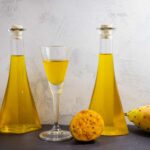 Zwei gläserne Karaffen und ein Glas mit gelblichem Kaktusfeigenlikör, umgeben von drei gelben Kaktusfeigen, eine halbiert.