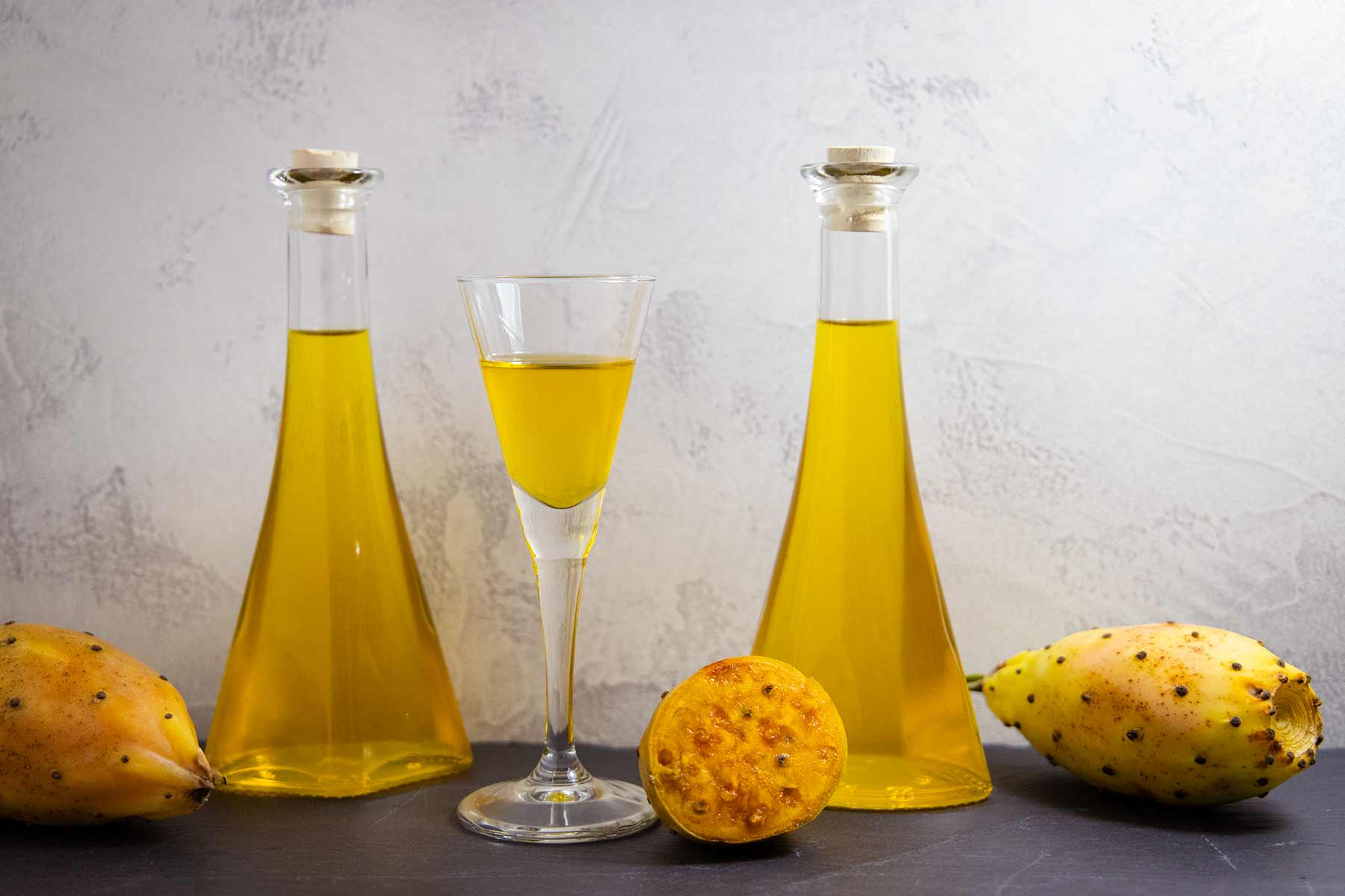 Zwei gläserne Karaffen und ein Glas mit gelblichem Kaktusfeigenlikör, umgeben von drei gelben Kaktusfeigen, eine halbiert.