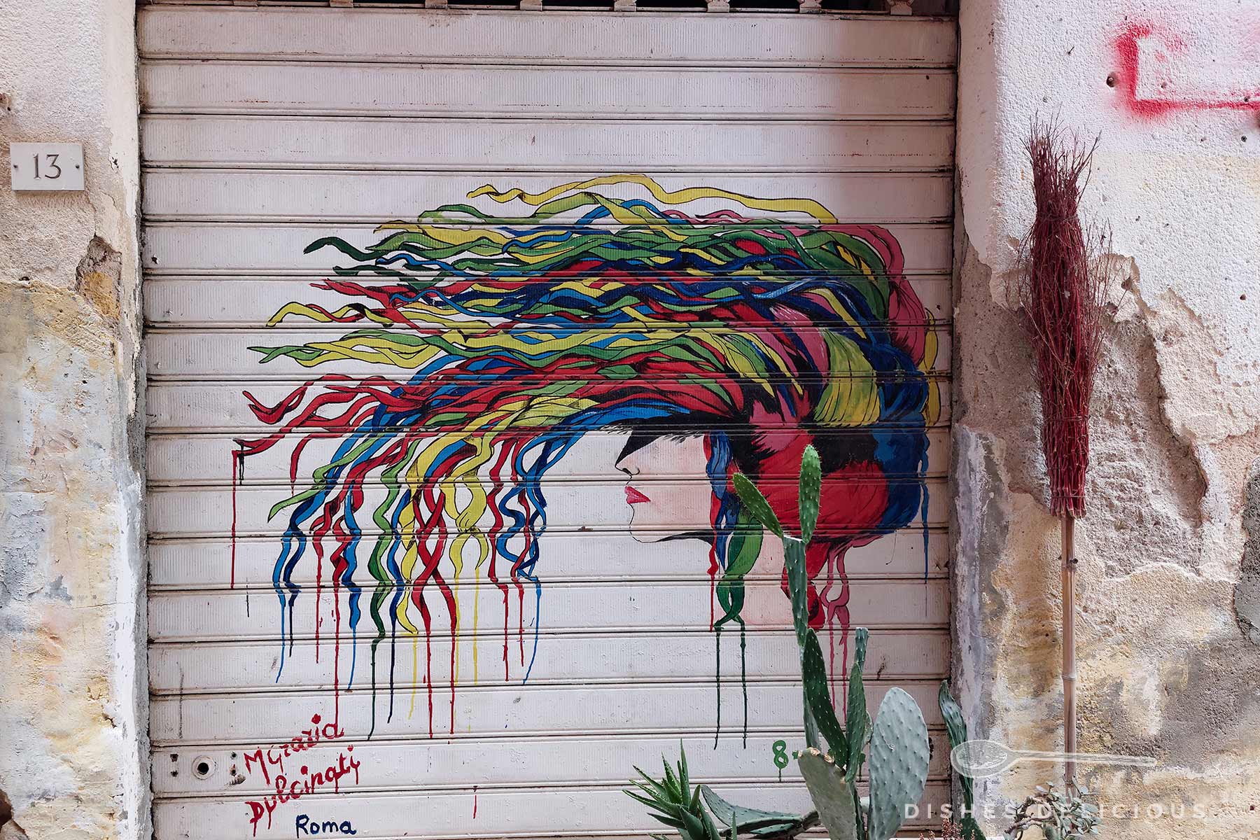 Bunte Wandmalerei einer Frau mit langem, wehendem Haar in Rot, Blau, Gelb und Grün auf einem Metalltor, links Hausnummer 13, rechts ein Bündel roter Zweige an der Wand.