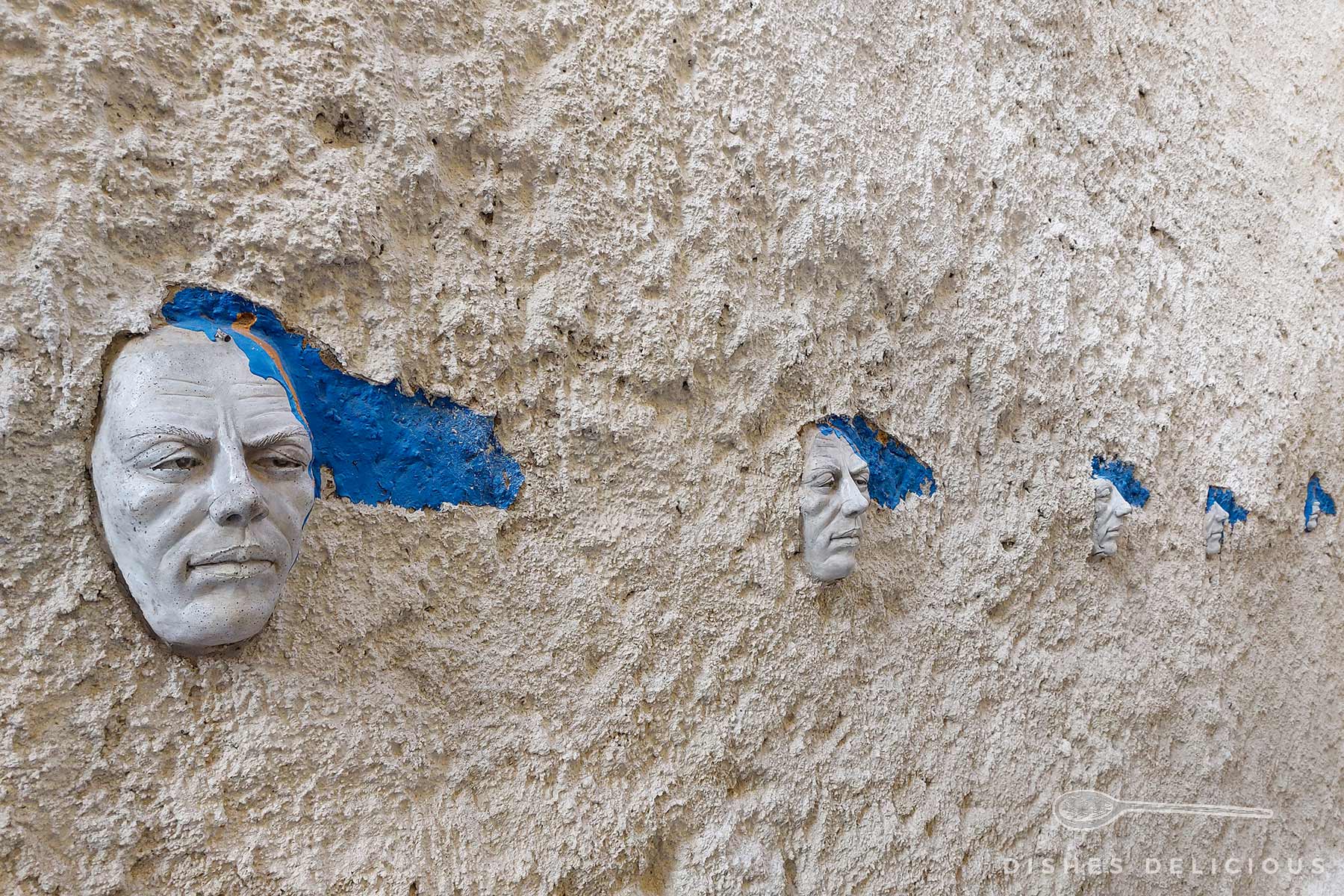 Mehrere kleine Gesichterskulpturen sind in eine raue Wand eingelassen, hinter ihnen sind blaue Farbflecken sichtbar.