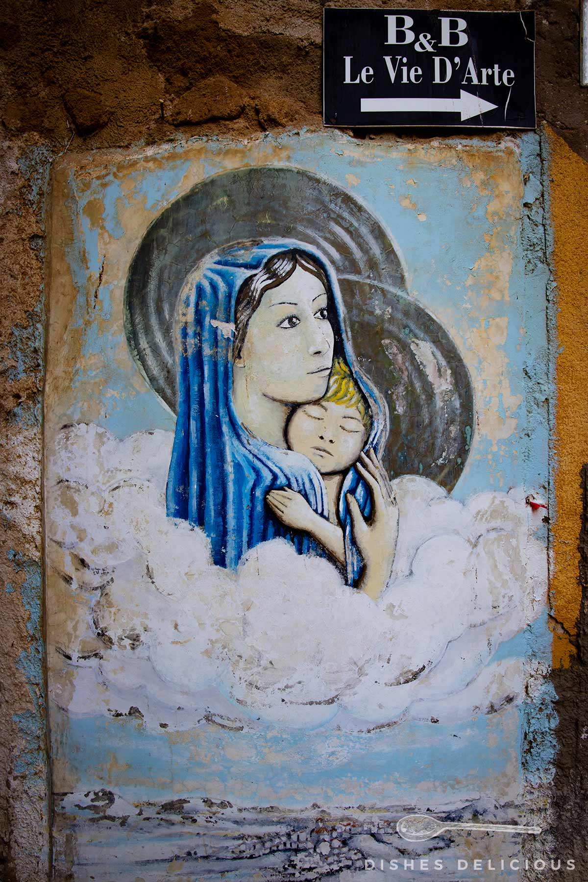 Wandbild einer Frau mit blauem Kopftuch und Heiligenschein, die ein Kind hält, umgeben von Wolken, darüber ein Schild mit der Aufschrift 'B&B Le Vie D'Arte' und einem Pfeil nach rechts.