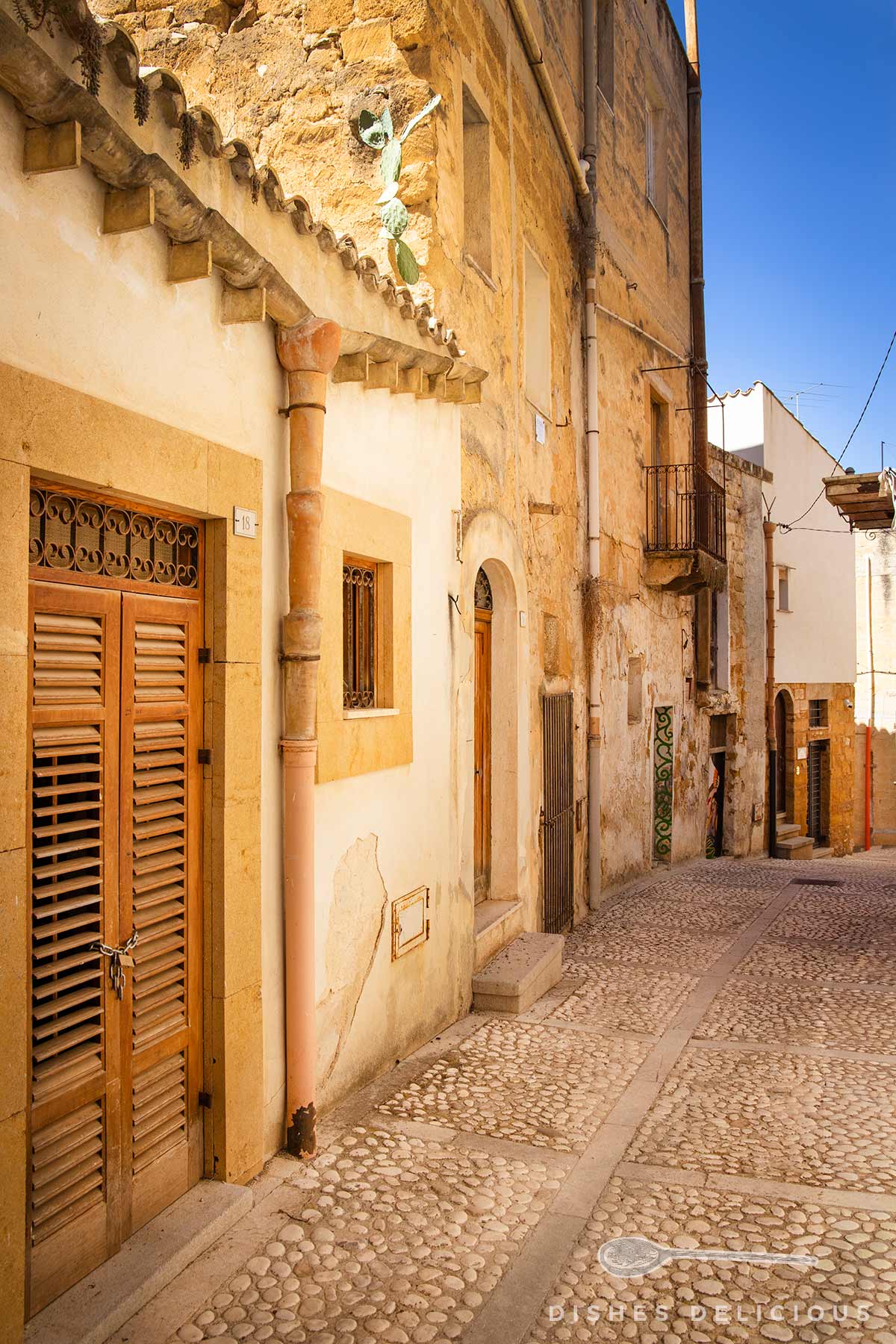 Sonnige Altstadtgasse mit Kopfsteinpflaster, gelblichen Häuserfassaden in Sambuca di Sicilia.