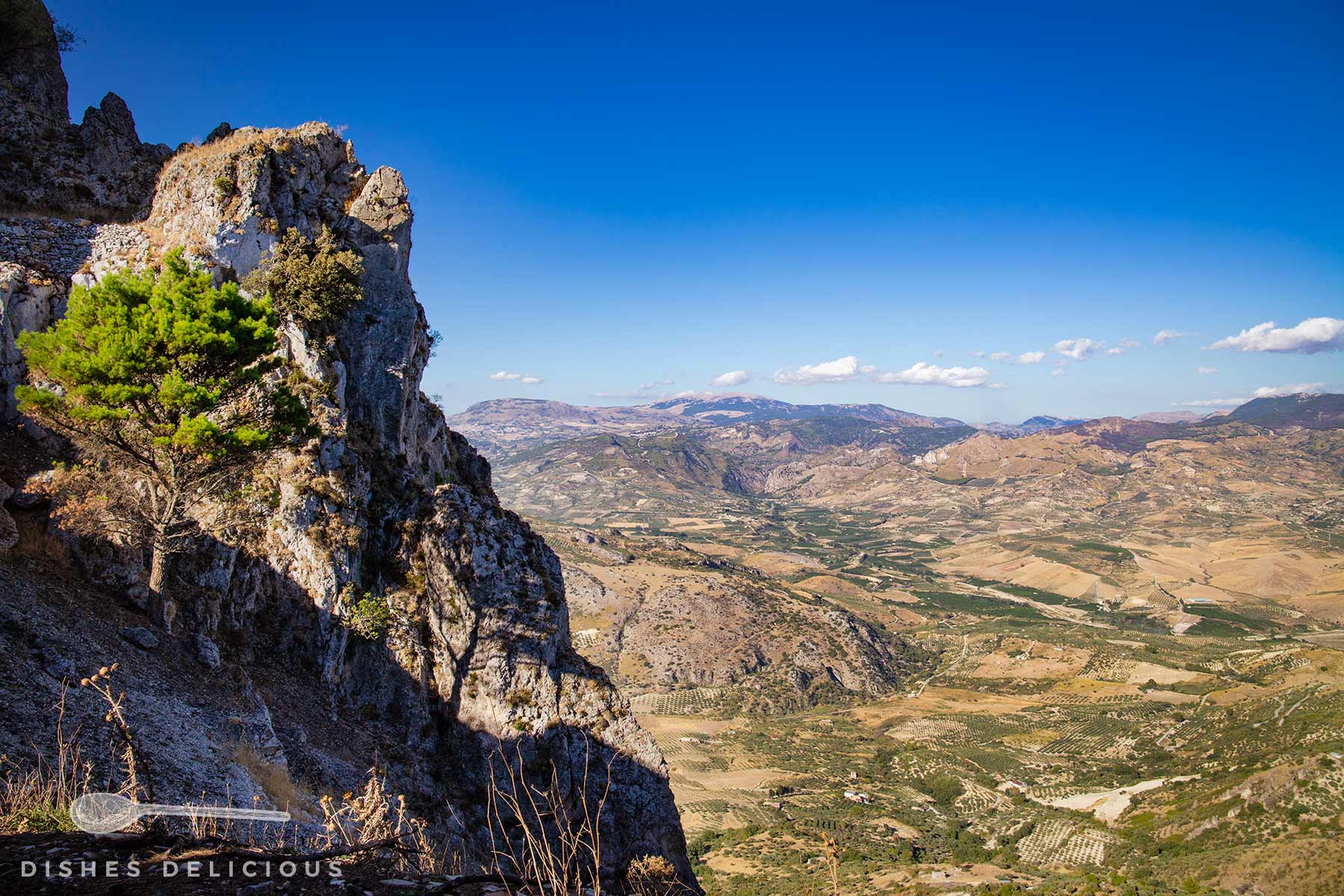Blick durch eine Felsöffnung auf ein weites, gebirgiges Tal unter blauem Himmel mit wenigen Wolken.