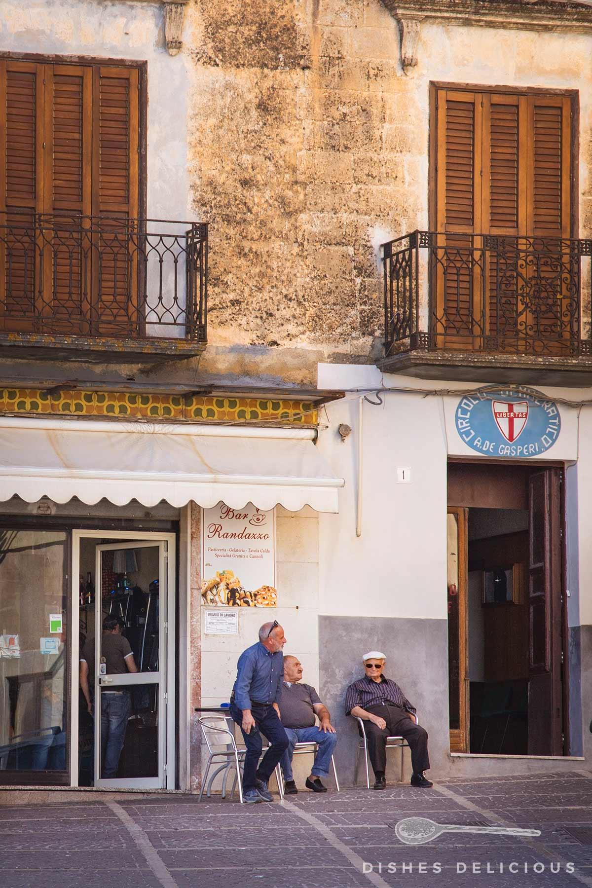 Caltabellotta, Sizilien: Drei Männer sitzen vor der Bar Randazzo, ein Gebäude mit zwei Balkonen und geschlossenen Fensterläden.