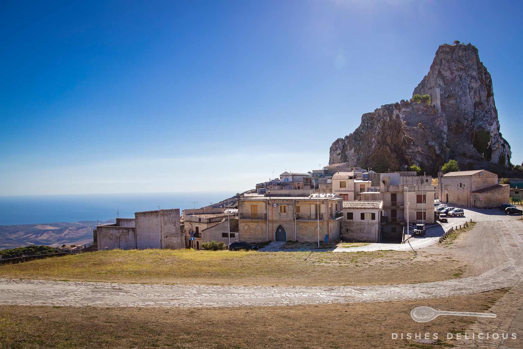 Häuser an der Bergspitze von Caltabellotta in Sizilien mit einem markanten Felsen im Hintergrund und Blick auf das Mittelmeer.