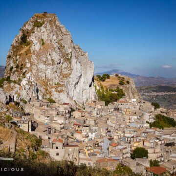 Sizilianisches Dorf Caltabellotta: Ein markanter Felsen überragt das dicht bebaute Dorf mit hellen Häusern. Im Hintergrund Berge.