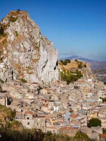 Sizilianisches Dorf Caltabellotta: Ein markanter Felsen überragt das dicht bebaute Dorf mit hellen Häusern. Im Hintergrund Berge.