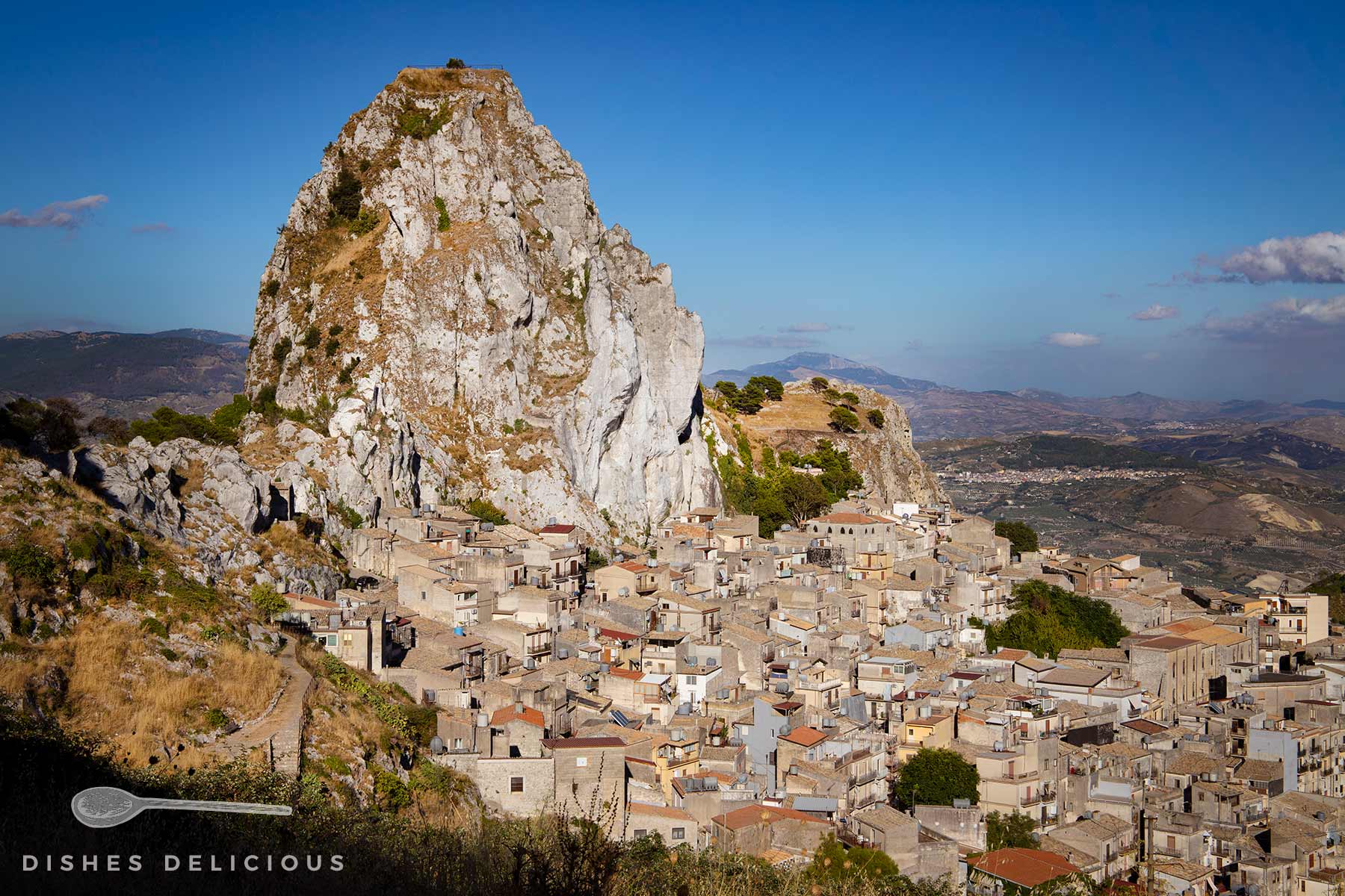 Sizilianisches Dorf Caltabellotta: Ein markanter Felsen überragt das dicht bebaute Dorf mit hellen Häusern. Im Hintergrund Berge.
