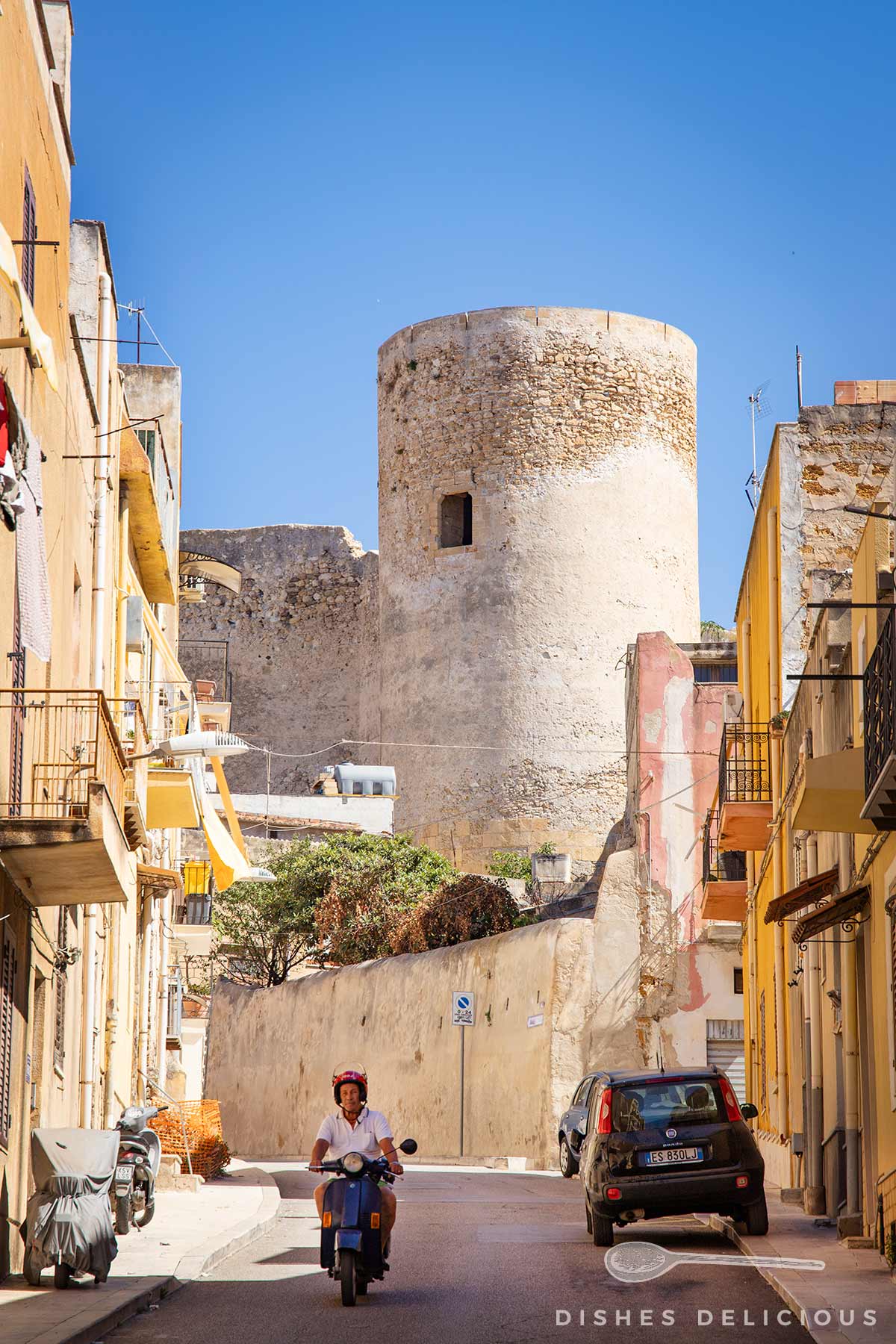 Runder Wehrturm des Castello Luna aus hellem Stein hinter schmaler Straße mit Häusern, parkendem Auto und Person auf Motorroller.