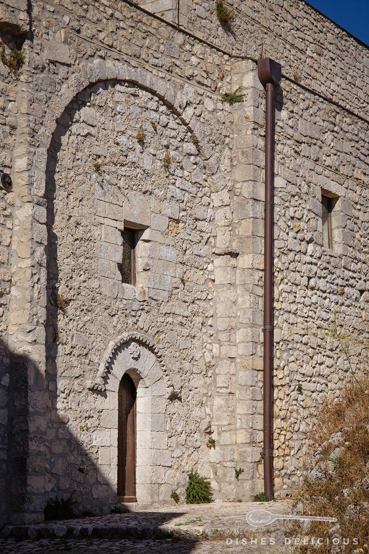 Fassade der Kathedrale in Caltabellotta aus grobem Stein mit kleinem, spitzbogigem Eingang und zwei kleinen Fenstern.