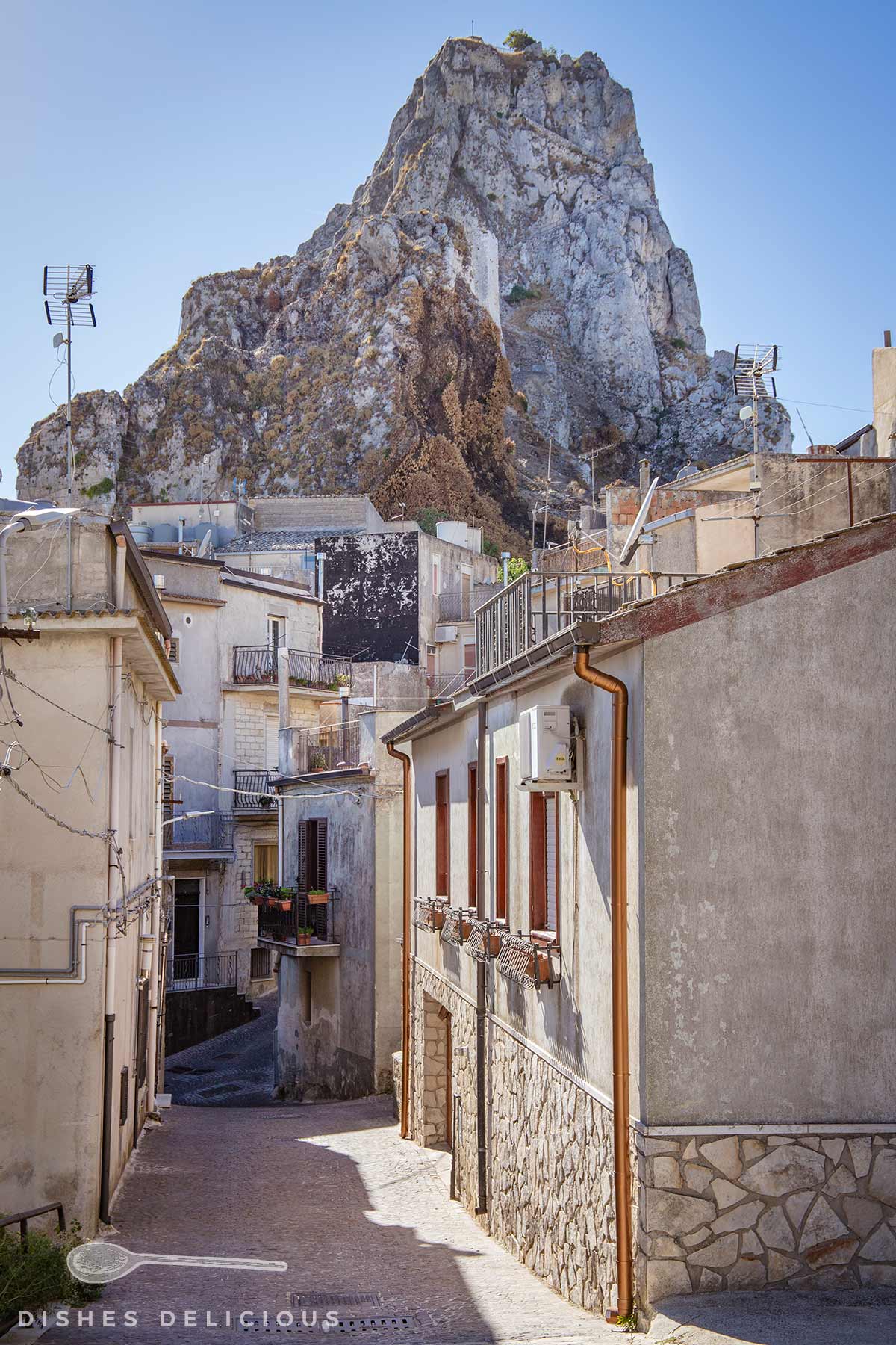 Enge Gasse mit hellen Häusern und Felsen im Hintergrund im historischen Zentrum von Caltabellotta.