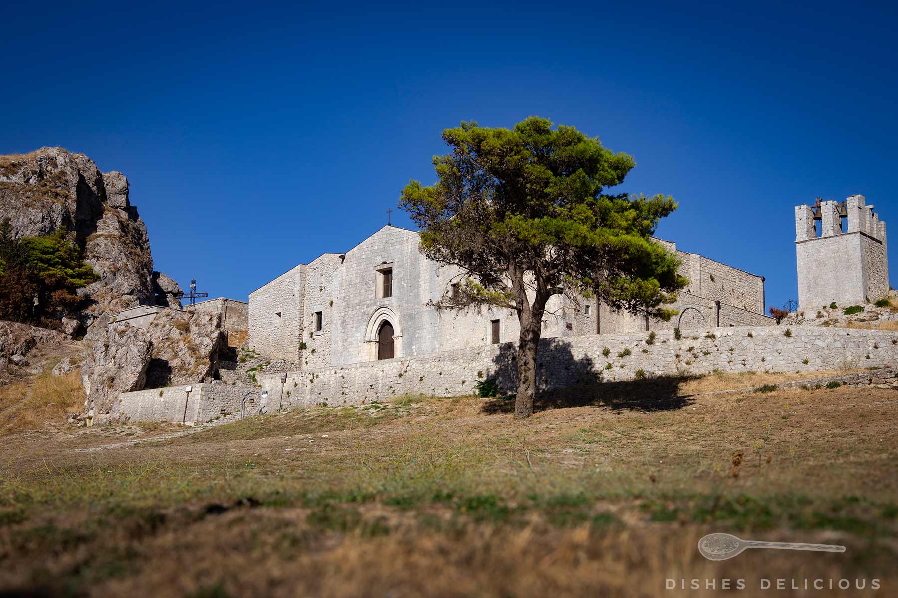 Chiesa Matrice unter blauem Himmel: Steinerne Kirche mit Rundbogenportal und angrenzendem Turm auf einem Hügel, davor ein Baum, links der Kirche Felsen.
