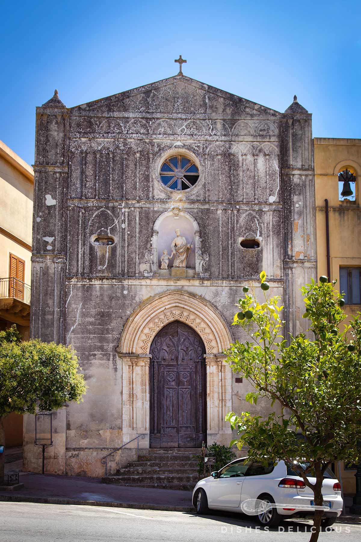 Fassade der Chiesa San Giuseppe in Sambuca mit rundem Fenster, Statue in Nische über einem großen Holztor, davor Bäume und geparktes Auto.