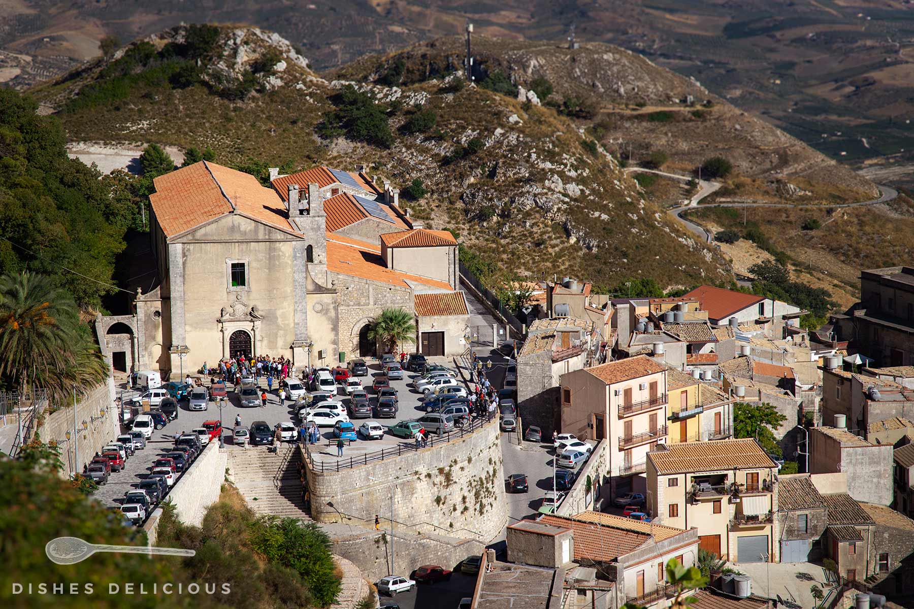 Blick vom Berg auf die Kirche Sant'Agostino in Caltabellotta, davor ein Parkplatz mit vielen Autos, umgeben von Häusern und Hügeln im Hintergrund.