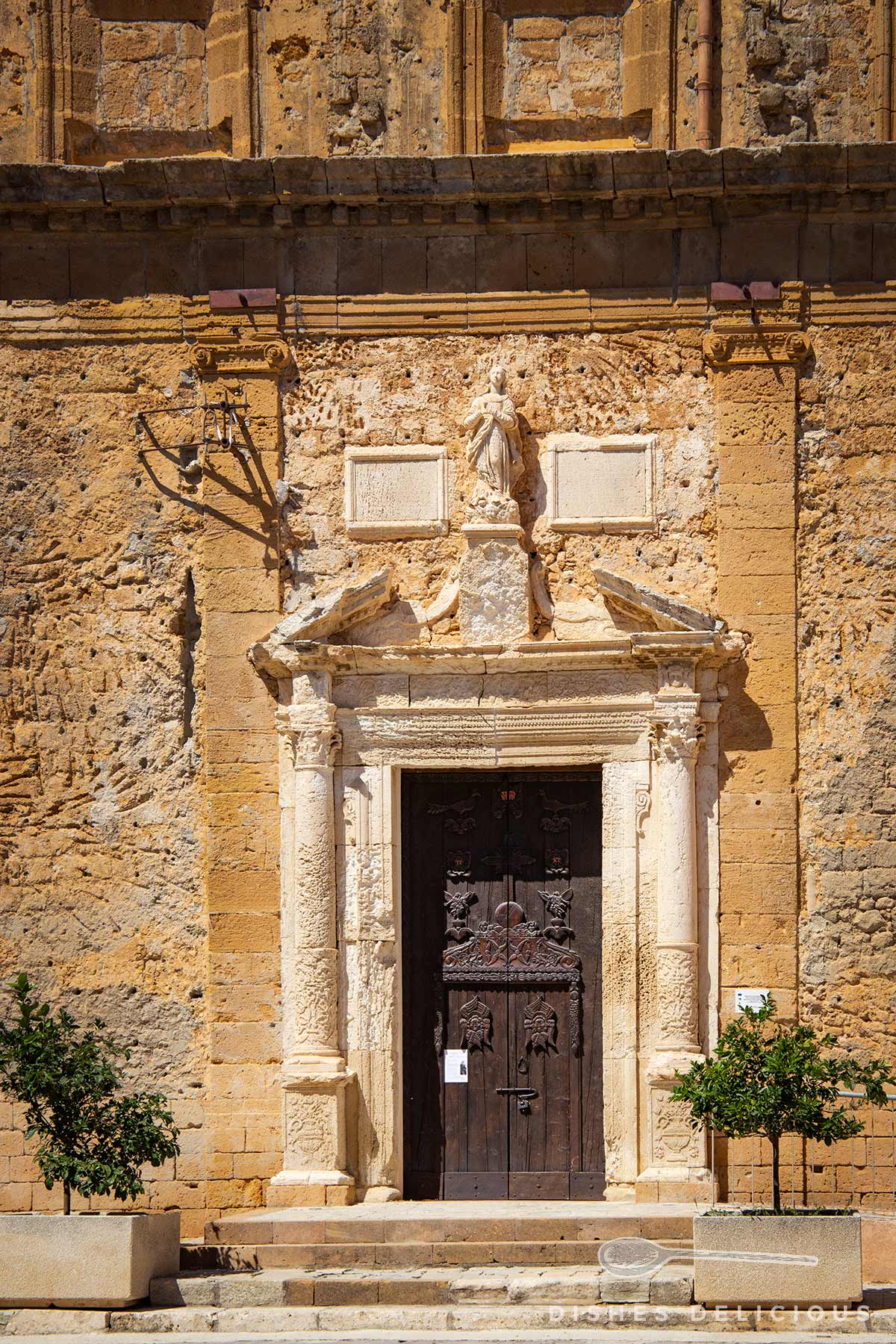 Eingang mit verziertem steinernem Portal und dunkler Holztür, darüber eine Statue und zwei rechteckige Tafeln, flankiert von zwei kleinen Bäumchen in Pflanzkästen.