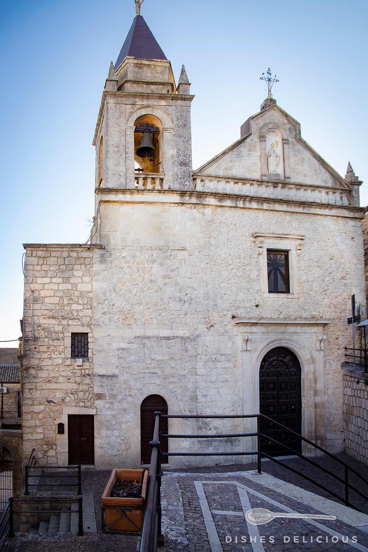 Kirche del Carmine mit Glockenturm und steinerner Fassade, rechteckige Fenster und dunkle Holztür, davor gepflasterter Platz mit einer Treppe und Geländer.