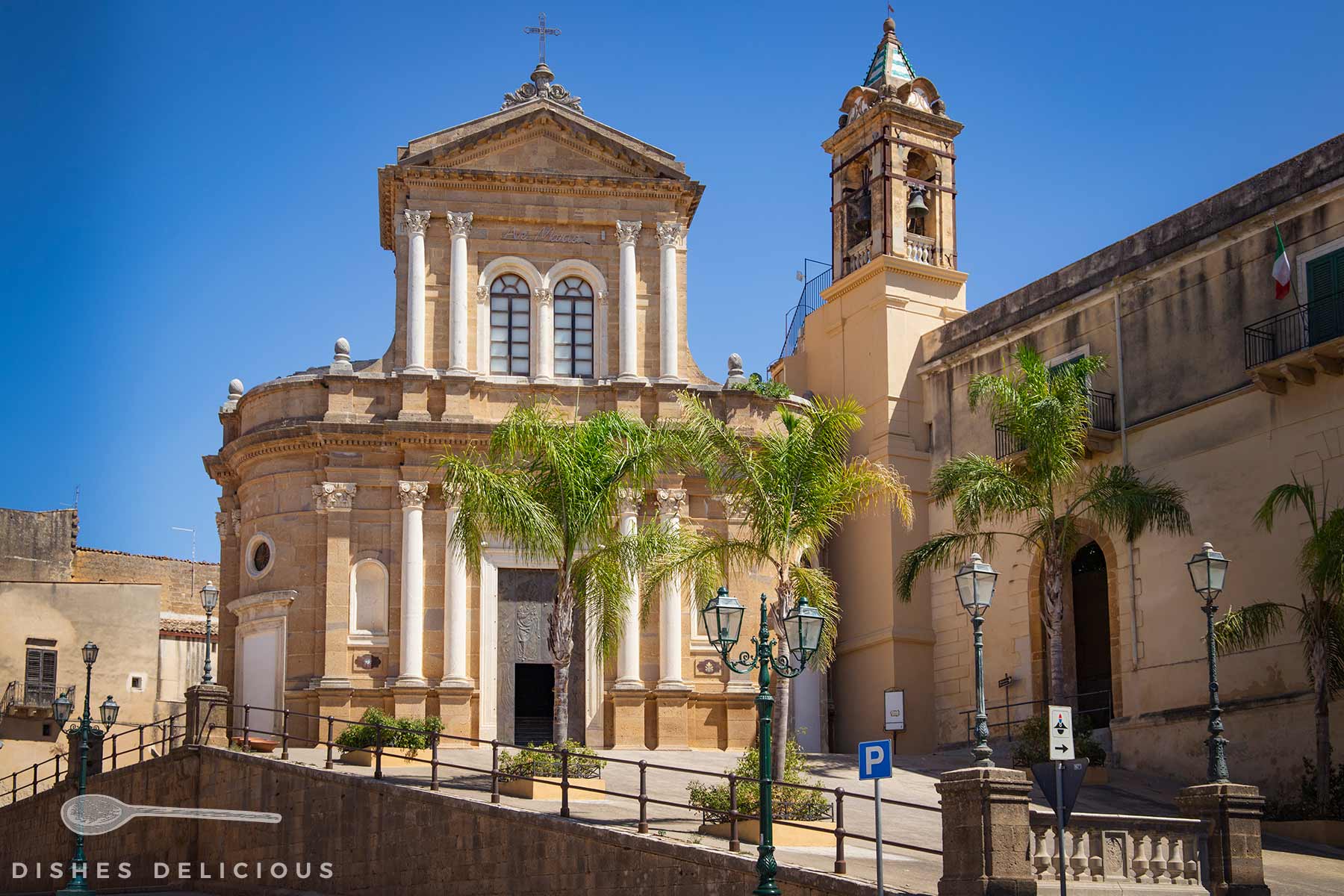 Barocke Carmine-Kirche mit Säulenfassade und Glockenturm, umgeben von Palmen und Straßenlaternen unter blauem Himmel.