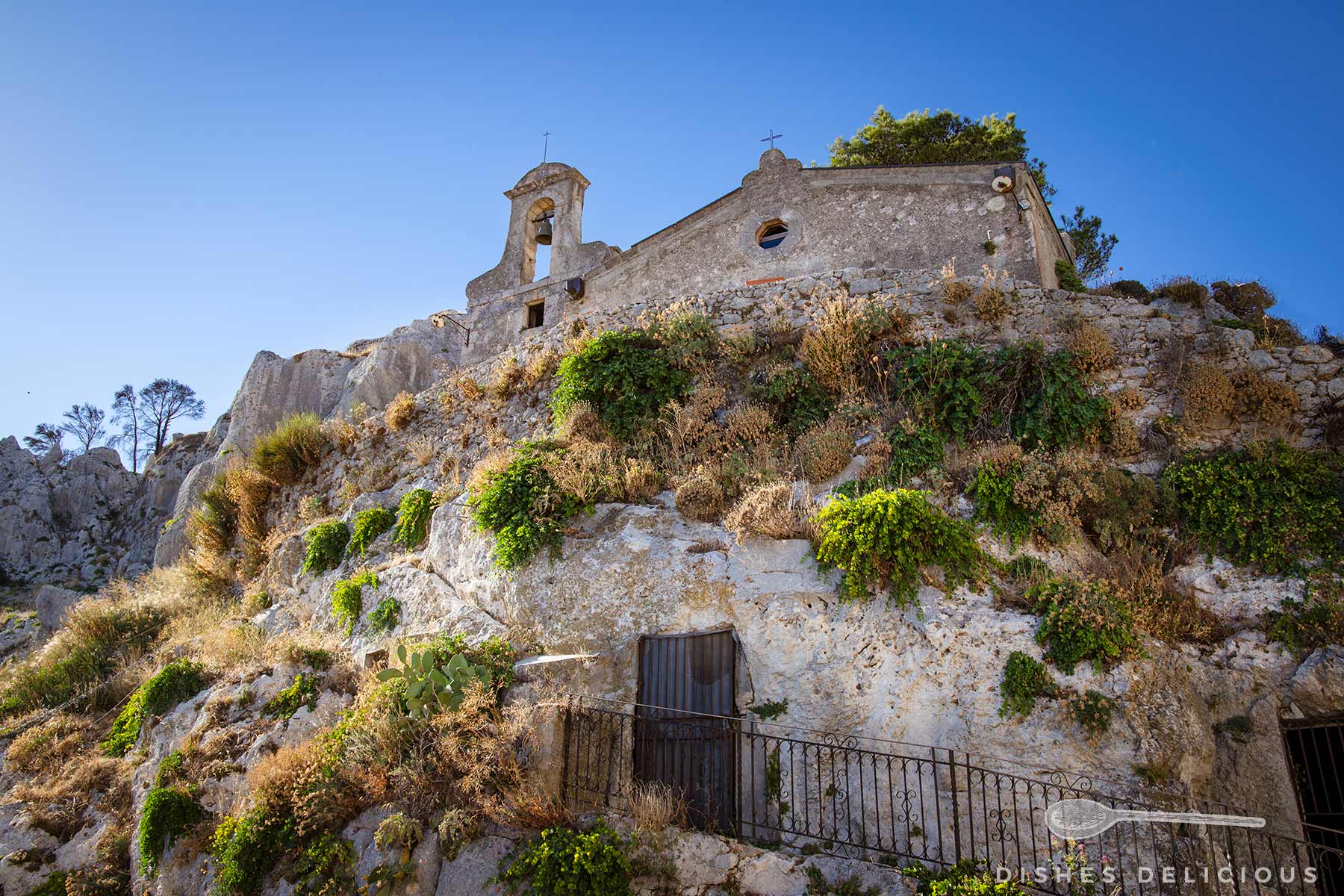 Chiesa della Pietà: Mit einem felsigen Hügel verwachsene Kirche mit Glockenturm, umgeben von trockenem Gras und Sträuchern unter blauem Himmel.