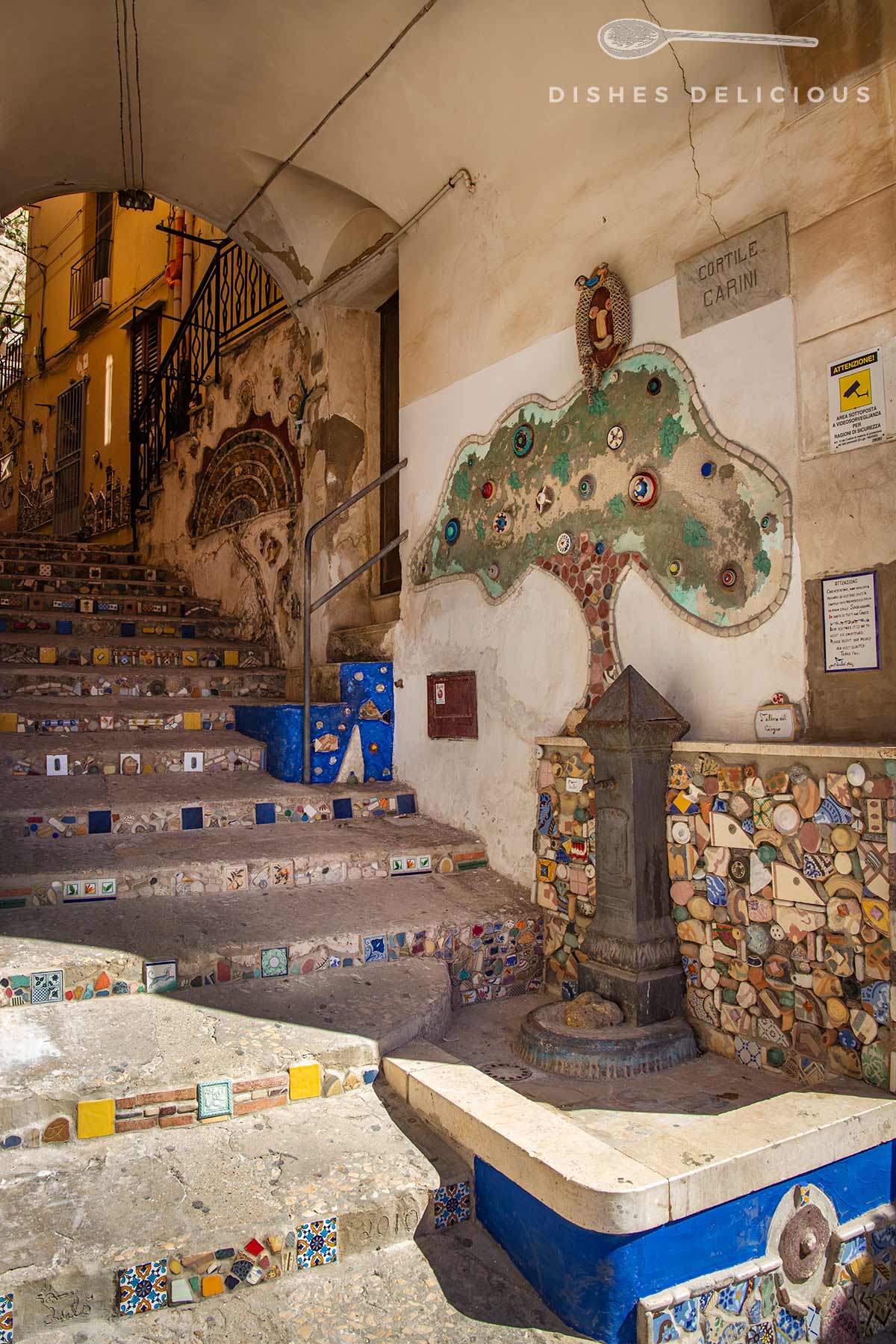 Treppenaufgang mit bunten Fliesen, Wandkarte von Cortile Carini und alter Wasserbrunnen unter einem Gewölbe.