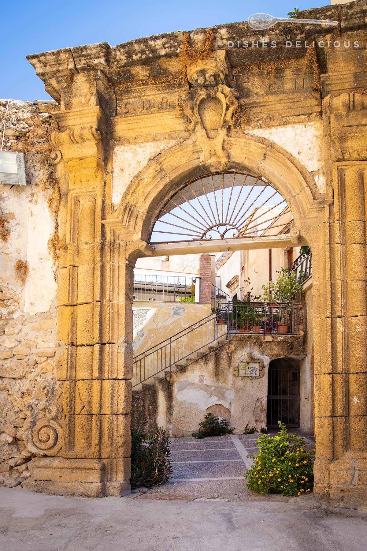 Sandsteinportal mit verziertem Bogen und schmiedeeisernem Gitter, dahinter Treppe und Pflanzen in einem historischen Gebäude.