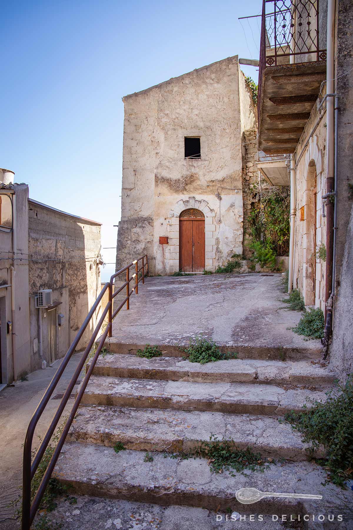 Schmale Gasse mit steinernen Stufen und altem, verwittertem Haus mit brauner Holztür und kleinem Fenster in Caltabellotta.