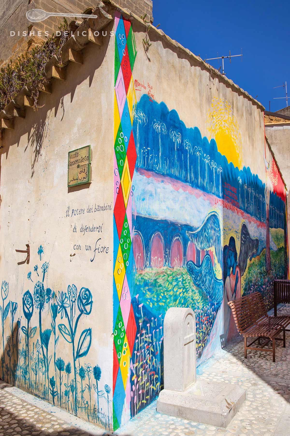 Bunte Wandmalerei an einem Haus mit Blumen, Brücken und Landschaftsmotiven, vor der Wand stehen zwei Holzbänke.