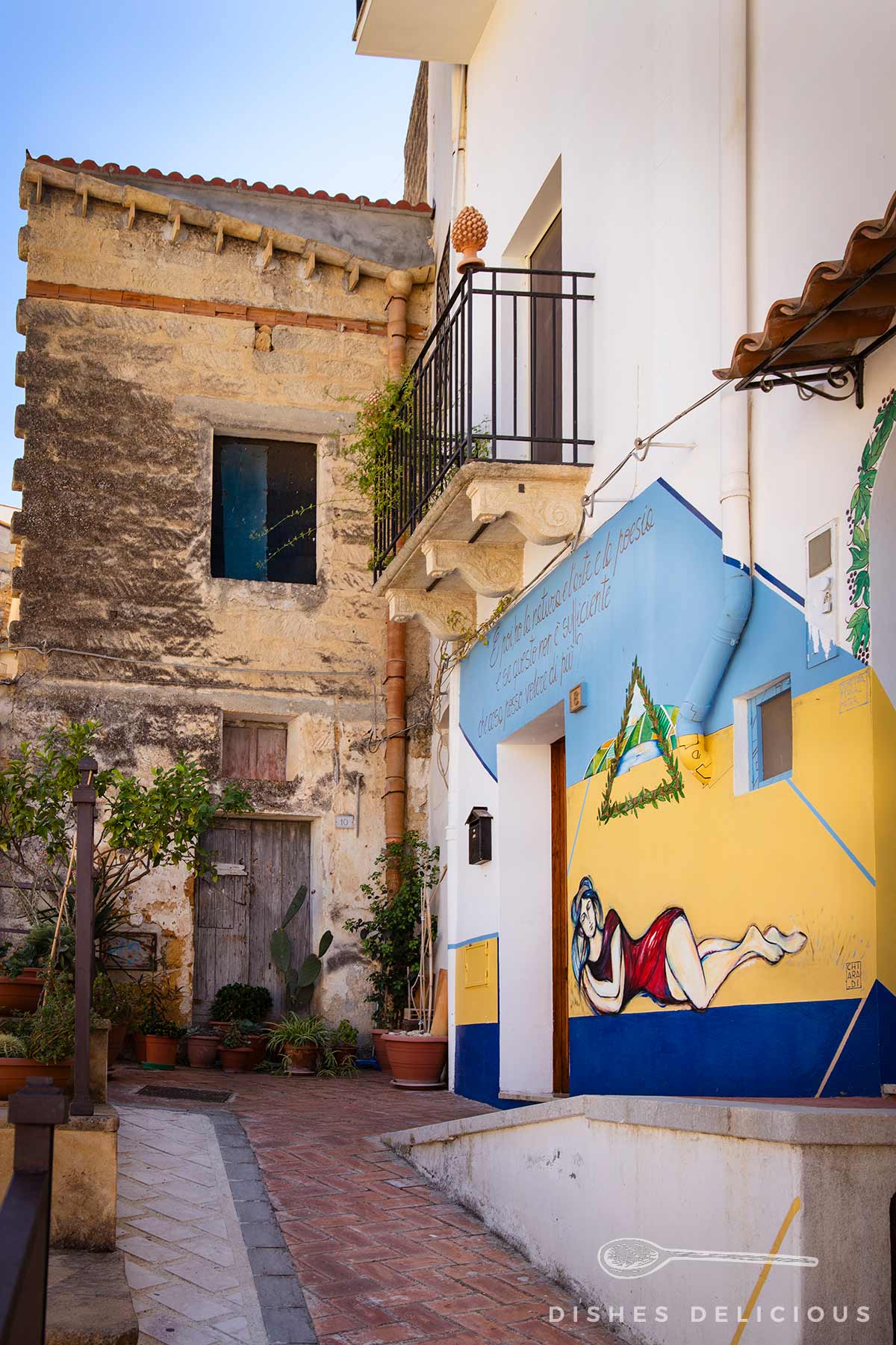 Steinhaus in der Altstadt von Sambuca. Hauswand mit Wandgemälde einer liegenden Frau in Rot und Weiß, Balkon mit schwarzem Geländer.