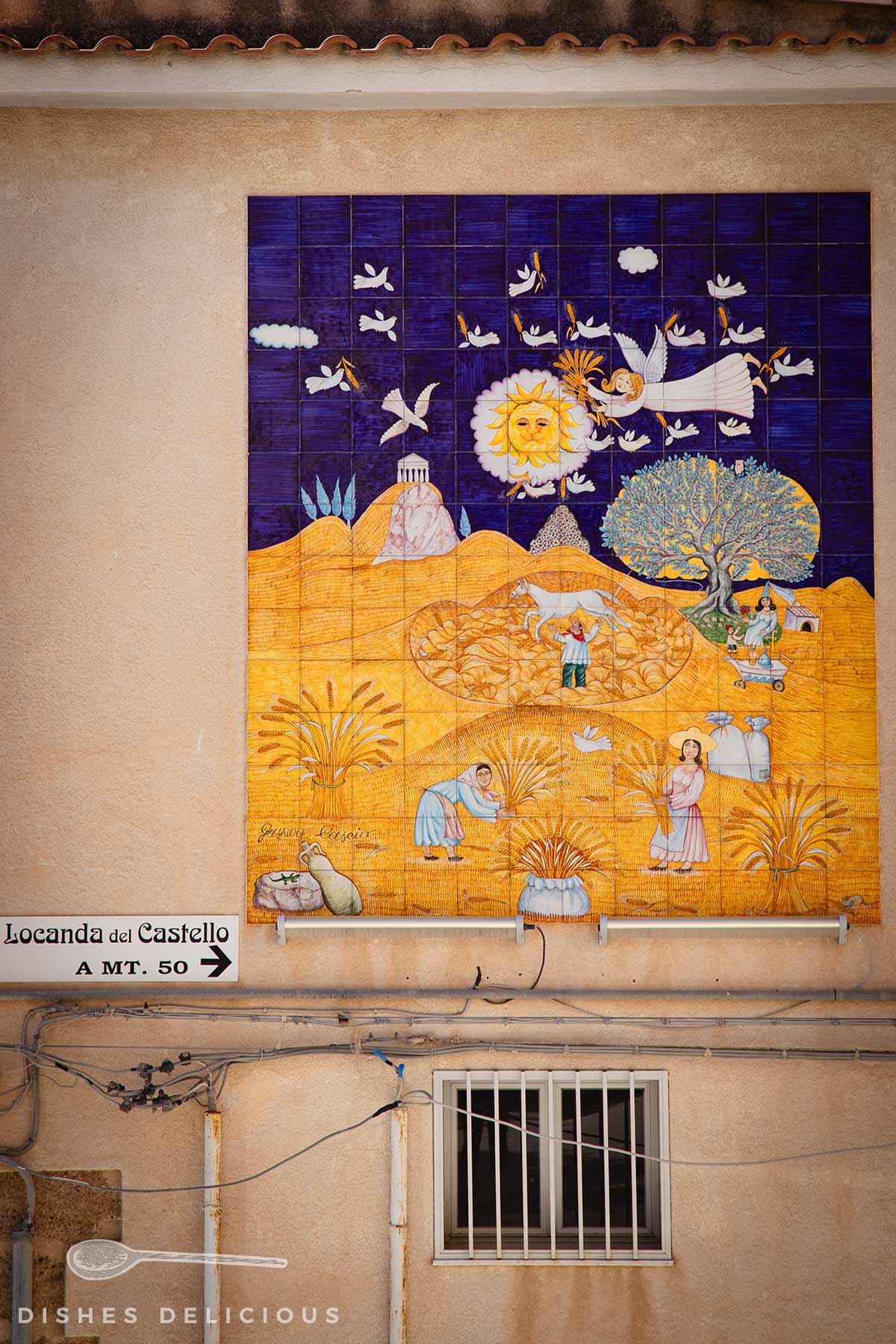 Keramikwandtafel mit Sonnenmotiv, Engel, Vögeln und Menschen in ländlicher Landschaft, darüber Dachziegel und darunter Fenster mit Gitter.