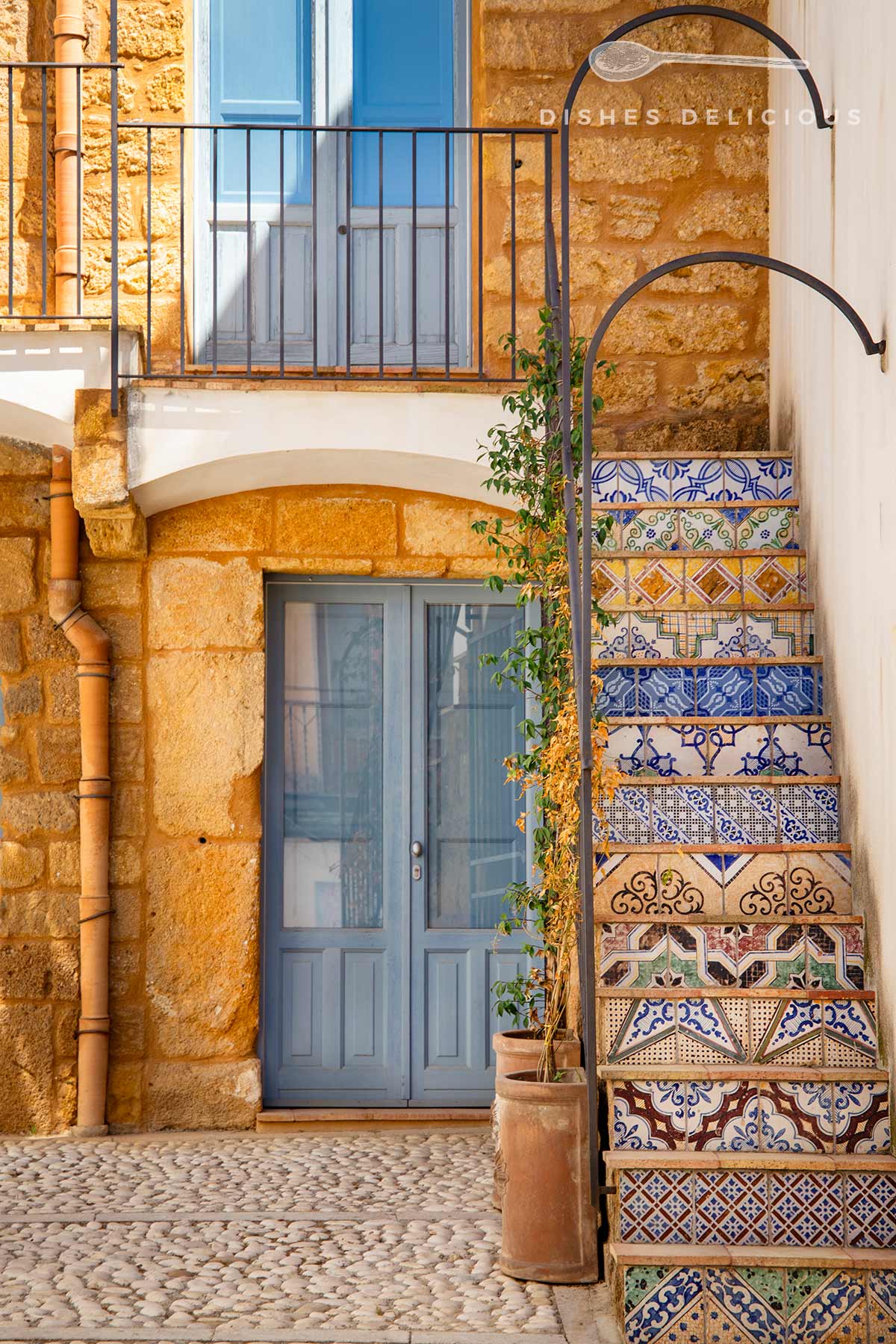 Keramiktreppe mit bunten, gemusterten Fliesen neben blau gestrichener Tür und Fenster in gelbem Sandsteingebäude.
