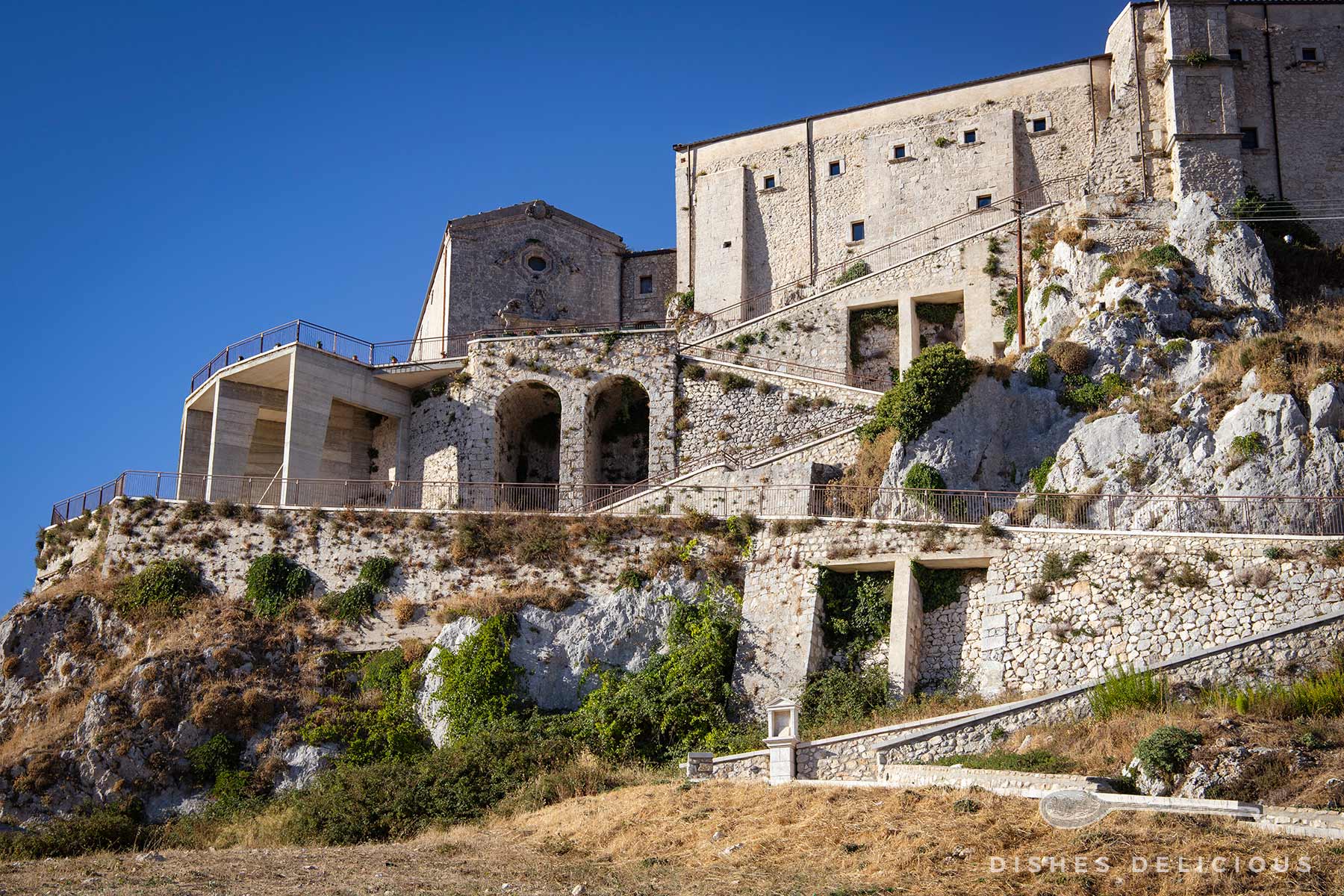 Kloster San Pellegrino auf felsigem Hang mit steinernen Mauern und Bögen unter blauem Himmel.