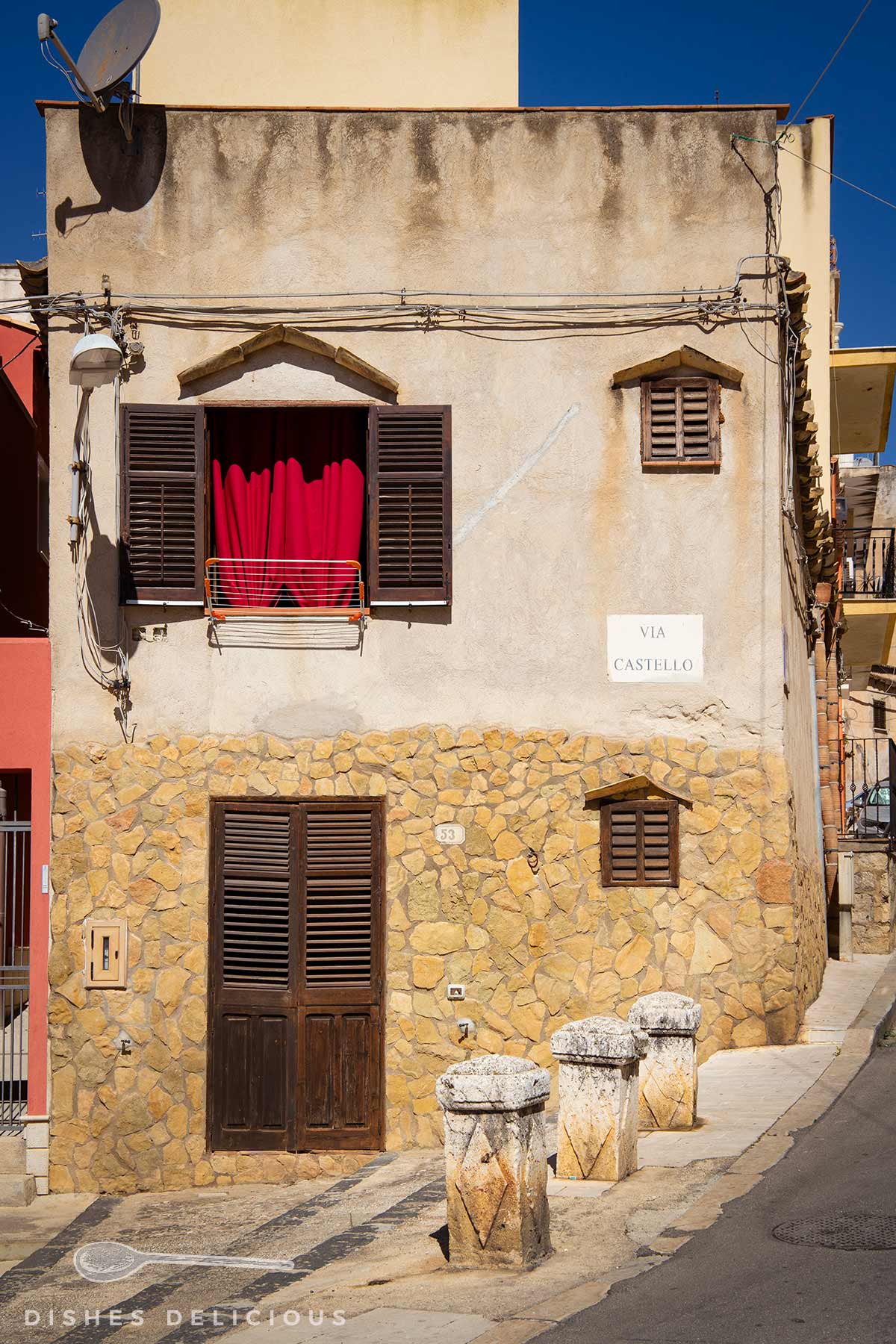 Fassade eines zweistöckigen Hauses mit steinverkleidetem Erdgeschoss, braunen Fensterläden, rotem Vorhang, Straßenschild 'Via Castello' und drei steinernen Pollern am Gehweg.