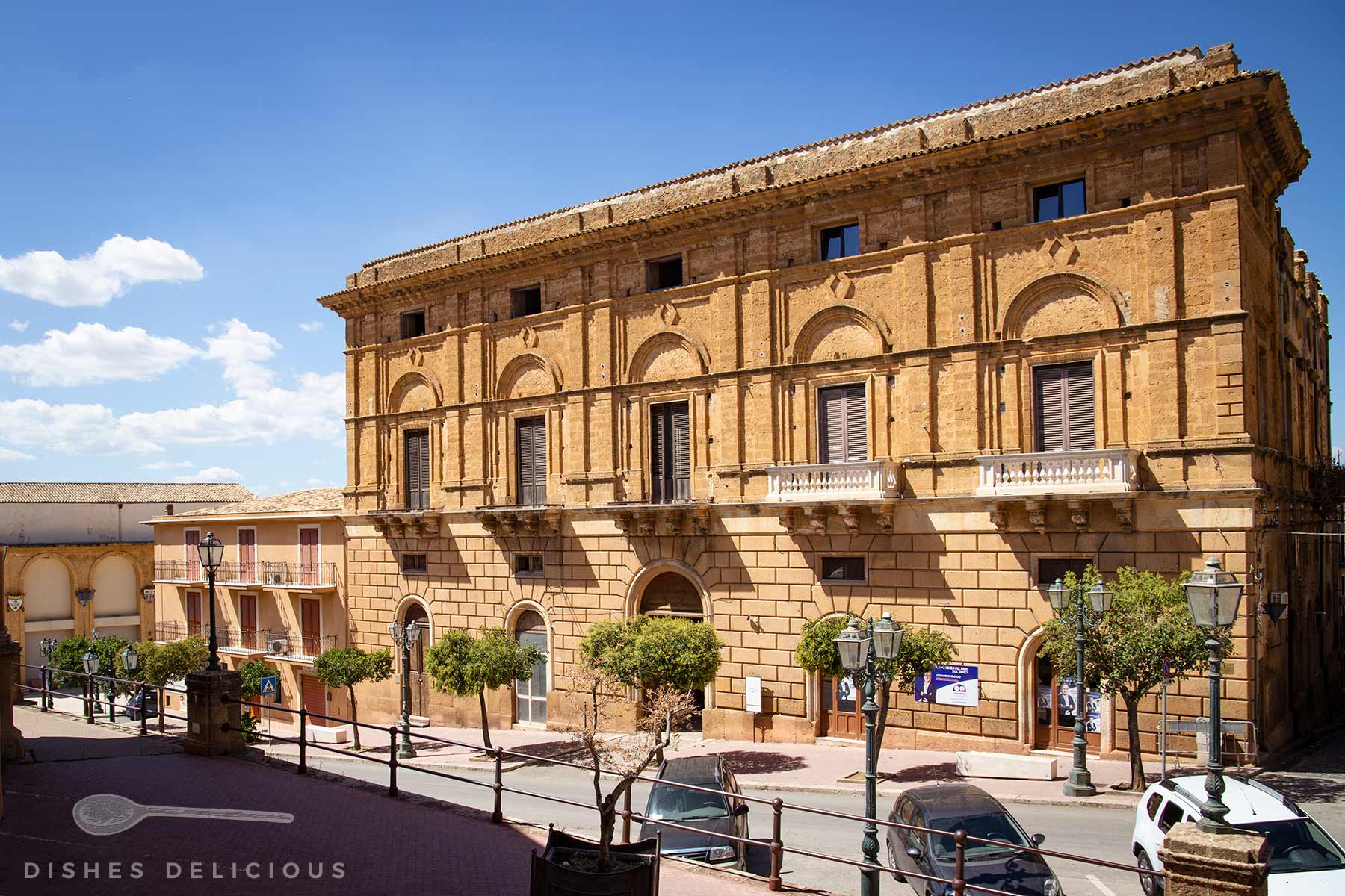 Fassadenansicht des Palazzo Ciaccio in Sambuca mit Verzierungen in Bogenform und Balkonen. An der Straße stehen Bäumen, sonniger Himmel.