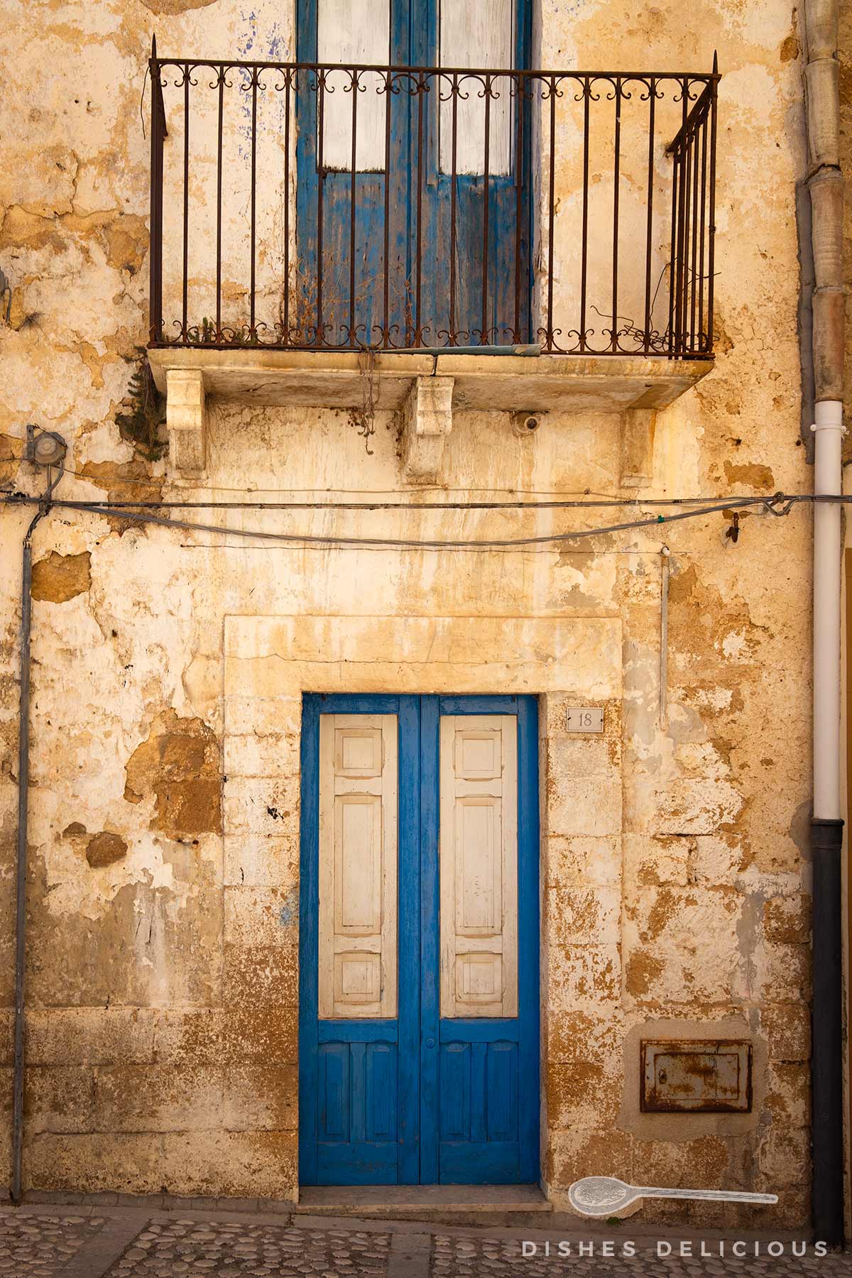 Fassade eines alten Hauses in der Altstadt von Sambuca di Sicilia mit blauer Doppeltür und kleinem Balkon mit schwarzem Geländer darüber.