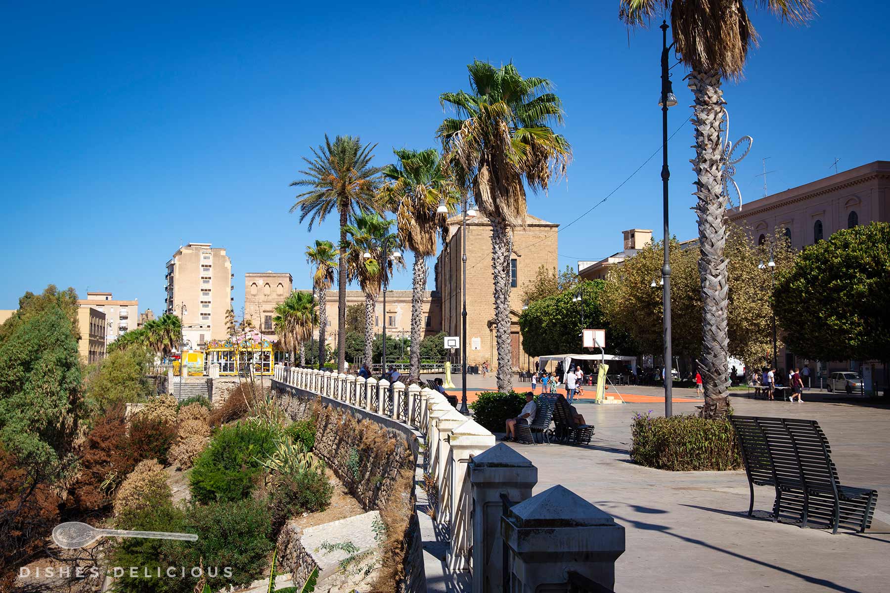 Piazza Scandaliato mit Palmen, Sitzbänken und Basketballkorb unter blauem Himmel und mit einer Kirche und anderen Gebäuden im Hintergrund.