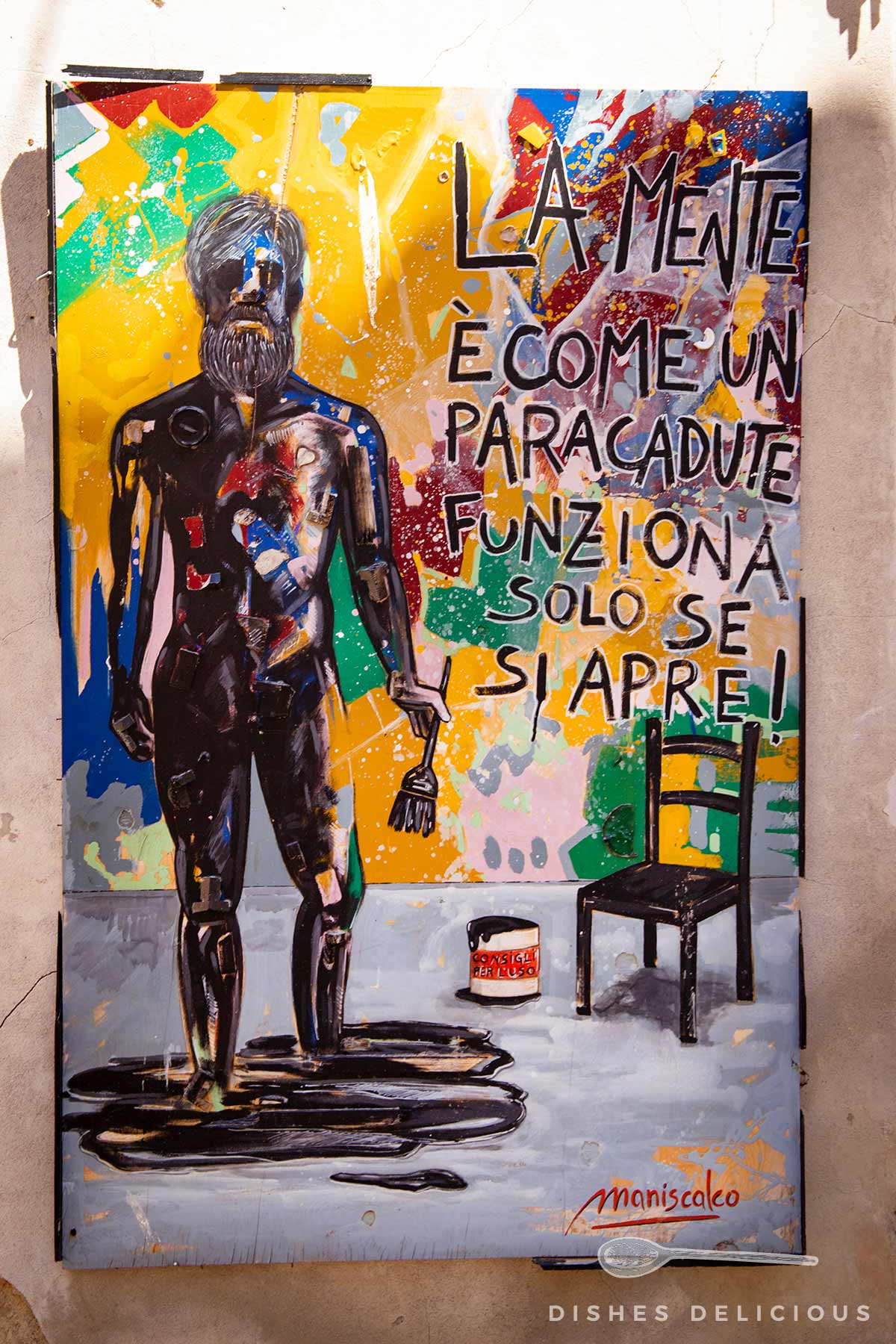 Bunte Street-Art mit männlicher Figur, die einen Pinsel hält, neben einem Stuhl und einem Farbeimer, dazu italienischer Text 'La mente è come un paracadute funziona solo se si apre!'