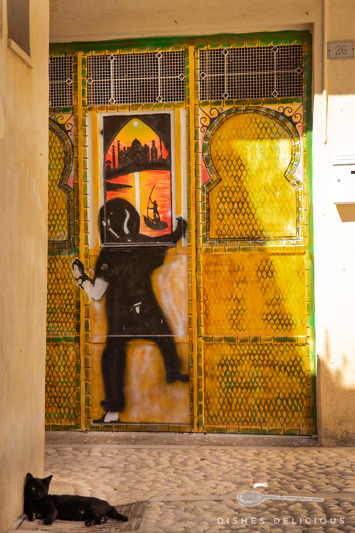 Gelb bemalte Metalltür mit Gittermuster und Street-Art-Malerei eines Mannes vor einer orientalischen Stadtlandschaft, schwarze Katze liegt auf Kopfsteinpflaster davor.
