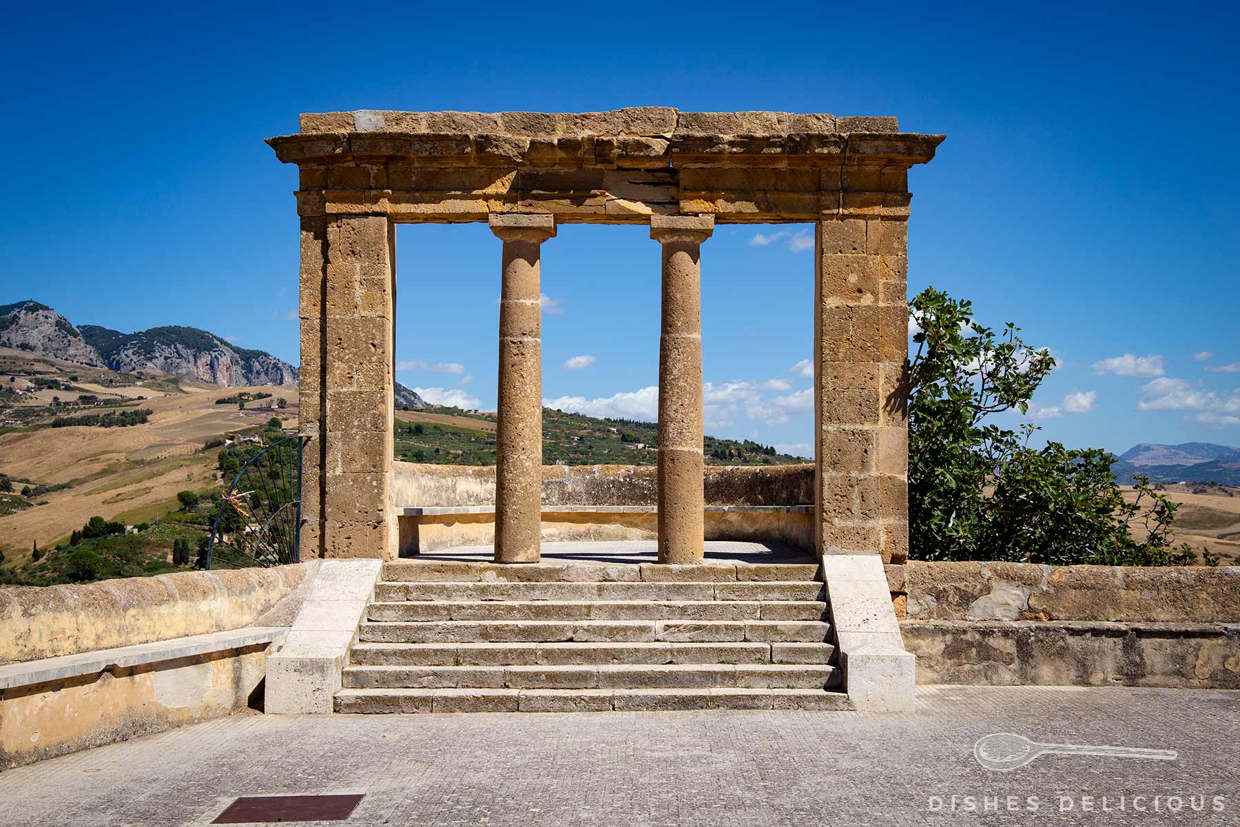 Terrazza Belvedere in Sambuca: Steintreppe führt zu antiker Säulenarchitektur mit drei Säulen, dahinter hügelige Landschaft und blauer Himmel mit Wolken.